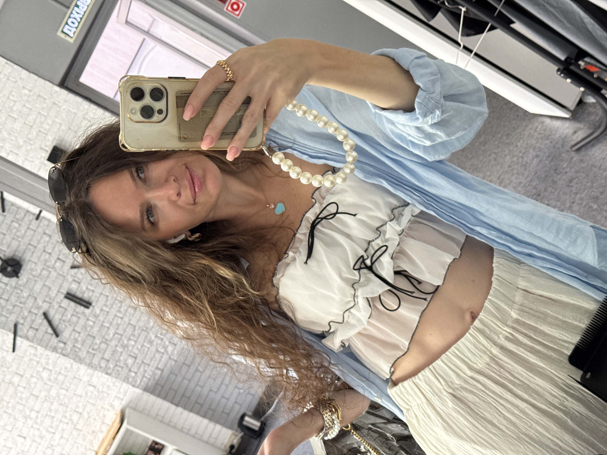 A woman with long, wavy brown hair and light blue eyes takes a selfie in a room that appears to be a salon or dressing room. She is wearing a white, off-the-shoulder top with ruffled edges and black ribbon ties, a light beige pleated skirt, and a light blue open-fronted shirt or jacket. She is holding a gold-colored smartphone with a clear case in her right hand, which has a pearl-beaded phone strap attached. Her left hand is partially visible, adorned with bracelets. She is also wearing a necklace with a turquoise heart-shaped pendant and a gold ring on her right index finger. Sunglasses are perched on top of her head. In the background, a large, modern clock with black hour and minute hands is mounted on a white brick wall. To the right, a partially visible door with a sign in Cyrillic that reads "ВЫХОД" (EXIT) is present. The floor is grey. The overall environment suggests a commercial establishment, possibly a beauty salon or boutique. There is no definitive indication of the city of Moscow, Russia, other than the implicit context provided.