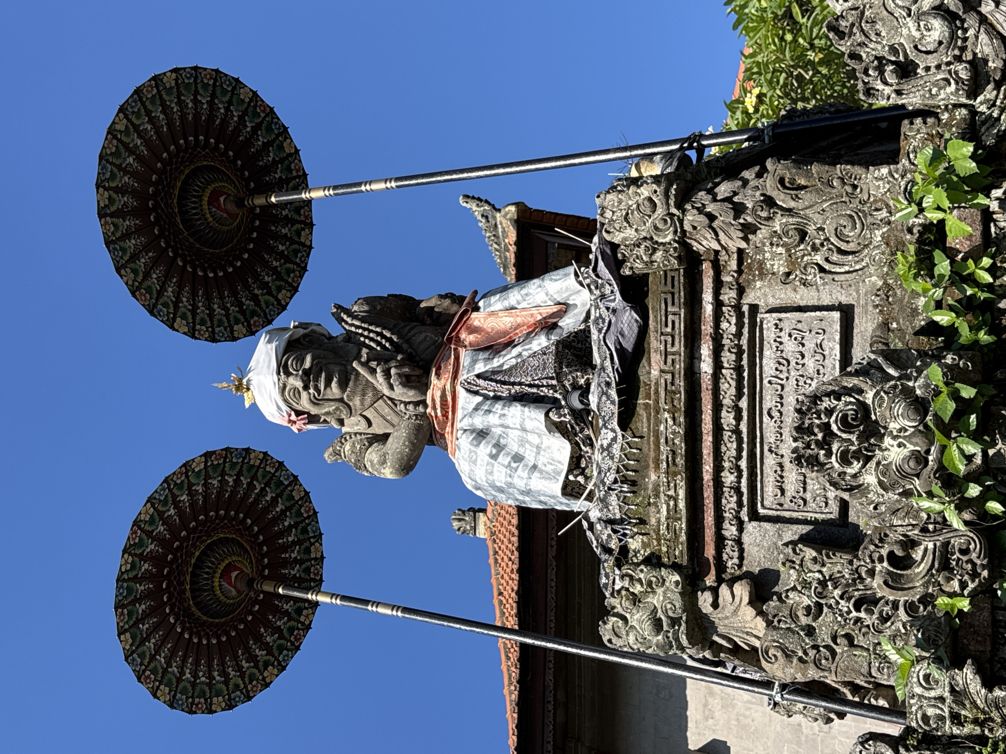 This image, captured in the Ubud District of Indonesia, showcases an ornate stone statue seated on a carved pedestal. Two decorative umbrellas, likely ceremonial, are positioned on either side of the statue, held aloft by dark poles. The statue is adorned with a white headdress and draped in patterned fabrics, including a prominent checkered cloth and a vibrant red sash. Its facial features suggest a smiling or laughing expression, and it appears to be holding an object in its hands, possibly a rosary or a small bundle. The pedestal itself is elaborately carved with intricate patterns and motifs, including what appear to be mythical creatures or gargoyles. Lush green foliage is visible growing around the base of the pedestal, and behind the statue, the terracotta roof tiles of a building are partially visible against a clear blue sky. The lighting suggests it is daytime, with the sun casting distinct shadows.
