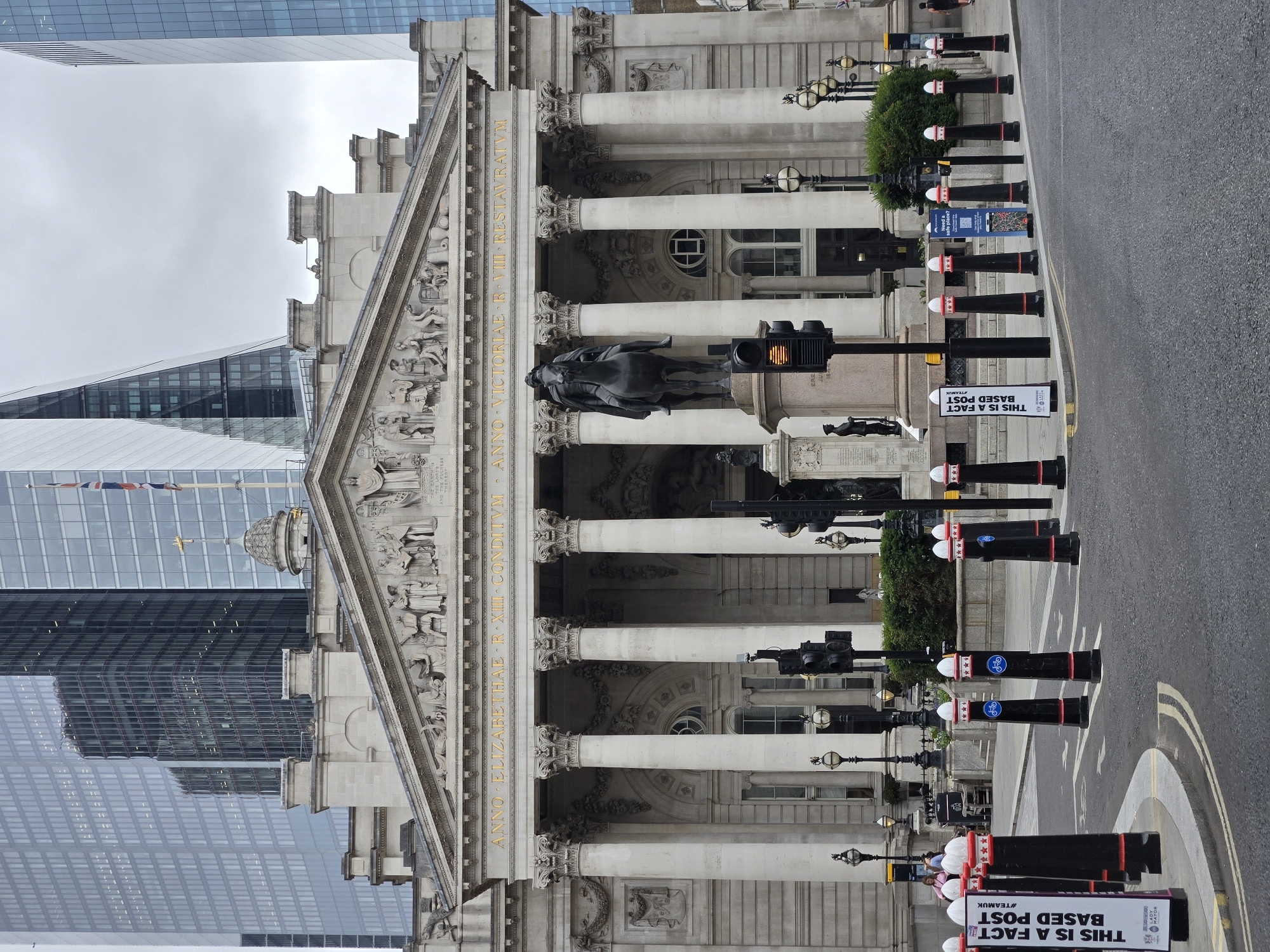 The image shows the Bank of England building in the City of London, United Kingdom, a grand neoclassical structure with a large pediment adorned with relief sculptures and prominent Corinthian columns. In front of the building stands a bronze equestrian statue. Modern skyscrapers dominate the background, contrasting with the historic architecture. The foreground features a street scene with traffic lights, bollards, and a pedestrian crossing. The sky is overcast, suggesting a cloudy day. Several bollards are visible, some with decorative elements and one with a sign that reads "THIS IS A FACT BASED POST #TEAMUK". A few people are present on the sidewalk and street, some walking, and others near a queue. The overall scene depicts a busy urban environment where historical and contemporary architecture coexist.