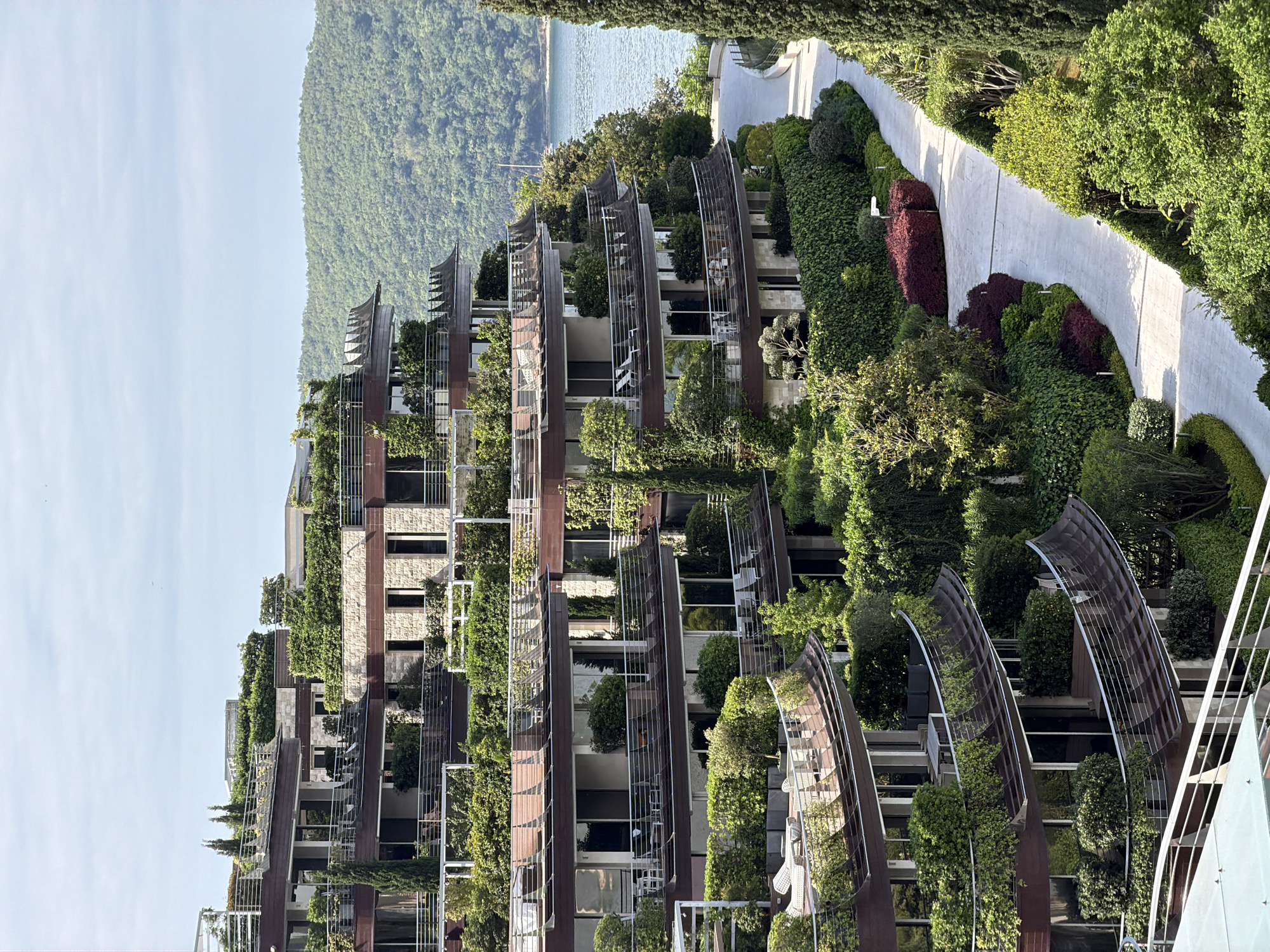 The image depicts a modern residential building complex in Budva, Montenegro, characterized by its extensive vertical landscaping. The building's facade features alternating sections of what appears to be stone or light-colored cladding and darker, possibly wooden or metal, elements. Lush greenery covers the balconies and other architectural features, integrating the building with the natural environment. The landscaping incorporates various plants, including trees, shrubs, and climbing vines.
The structure includes curved, metal-framed sunshades or architectural elements that extend outward from the facade. The building is set against a backdrop of wooded hills and a glimpse of the Adriatic Sea. To the right of the building is a winding, light-colored pathway or sidewalk lined with manicured hedges and gardens, including a patch of dark-red bushes. No people are visible in the image.