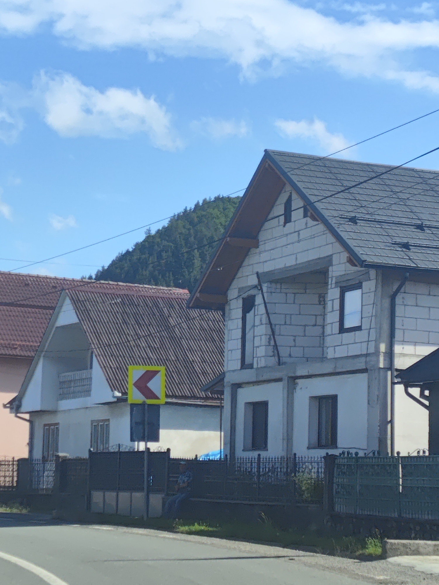 The image shows a street scene in Leordina, Romania, with several houses visible. The foreground features a paved road with white lane markings. Behind the road, there is a dark metal fence and grass. Several houses are depicted: one on the left with a pinkish facade and a brown tiled roof, a white house with a corrugated roof in the center, and a house under construction on the right, made of white cinder blocks with a dark tiled roof. A yellow and red chevron traffic sign indicating a curve to the left is mounted on a pole in front of the central white house. A person is seated on a bench behind the fence, facing away from the camera, wearing a plaid shirt and blue jeans. The background consists of a tree-covered hillside under a bright blue sky with scattered white clouds. Power lines are visible crisscrossing the sky.