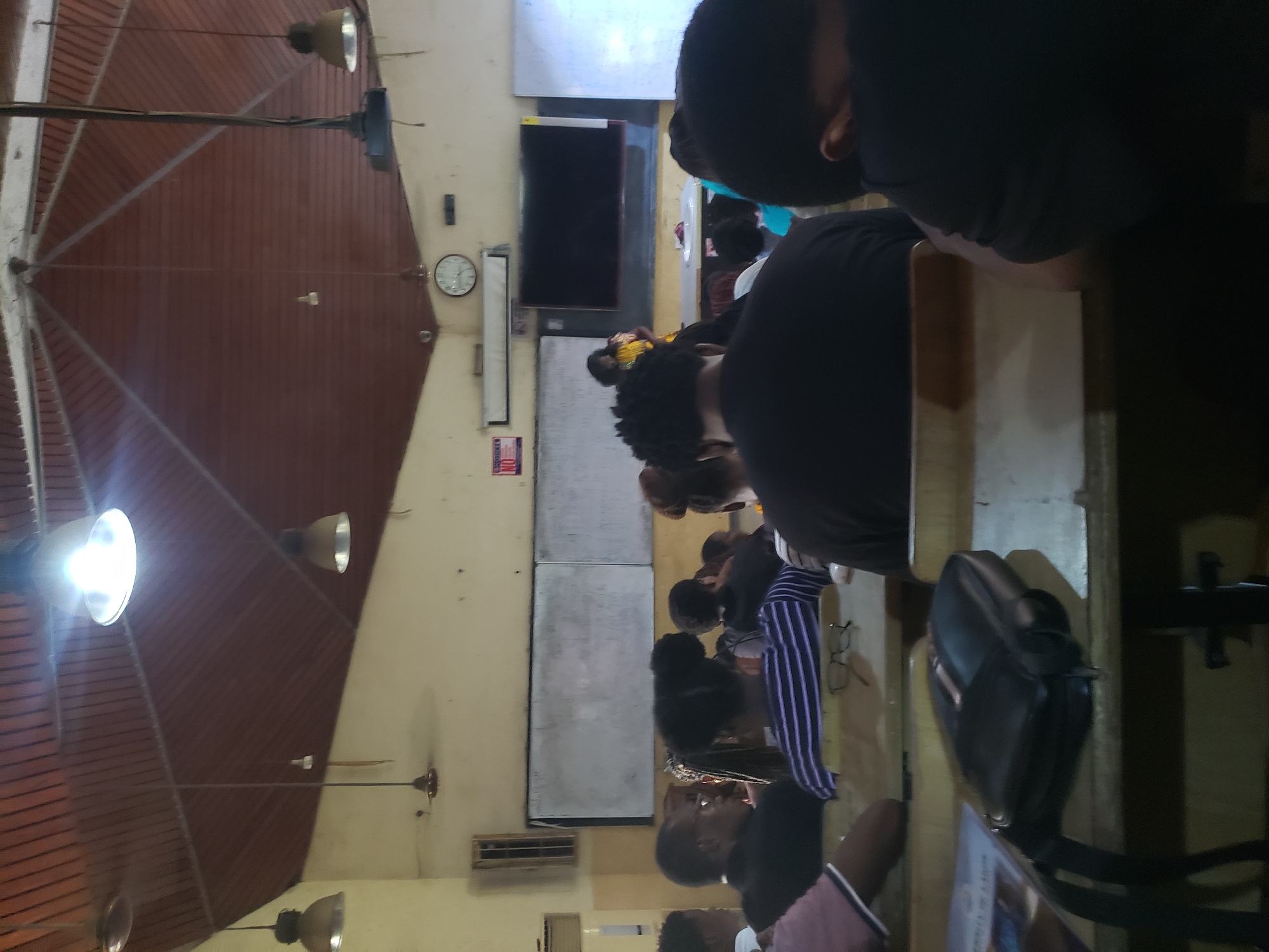 The image depicts an indoor scene, likely a classroom or lecture hall, with a group of people seated at desks, facing a presenter. The ceiling is constructed of wooden planks with exposed beams and features several industrial-style light fixtures. A large, dark screen is mounted on the wall behind the presenter. Two whiteboards are also visible on the wall. The overall atmosphere appears to be that of an educational setting. The image is likely taken in Iganmu, Nigeria, given the context of the prompt.