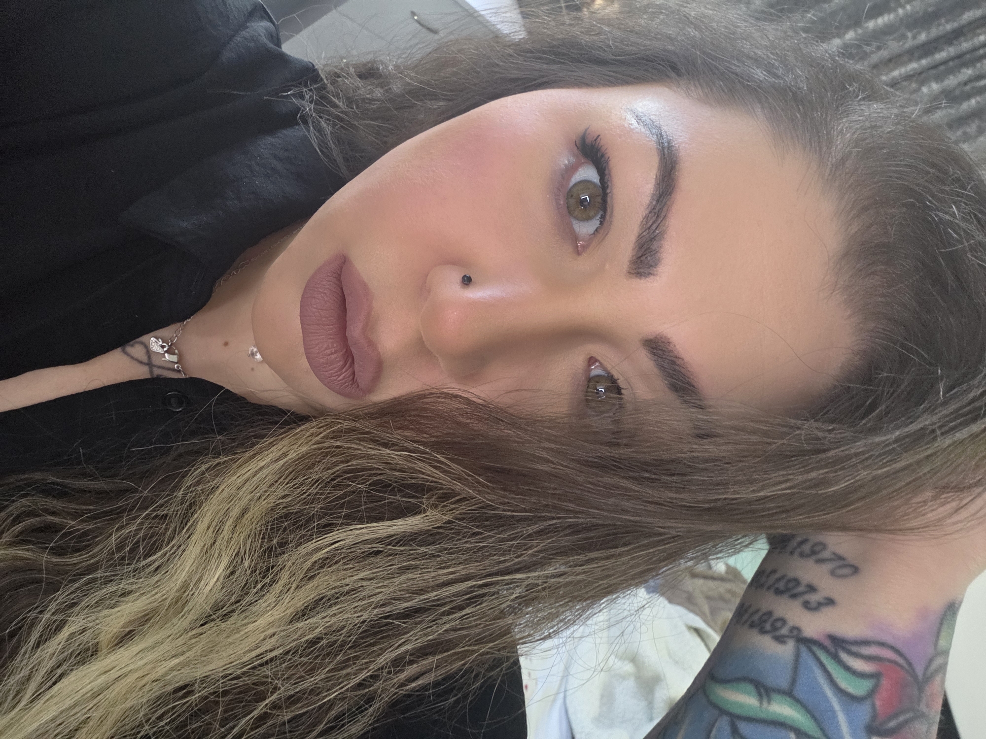A close-up, angled portrait shows a person with fair skin, hazel eyes, and dark brown hair with blonde highlights framing their face. They wear a dark colored shirt or top. The person has well-defined eyebrows, full lips with a mauve lipstick, and a small stud piercing on the left side of their nose. A delicate silver necklace with a charm is visible on their neck. The person's left arm and wrist is visible, featuring a colorful tattoo with dates "1970, 1973, 1992." The background appears to be a light, neutral color, possibly a wall or ceiling. There is nothing in the image to suggest the location is the city of London, United Kingdom.