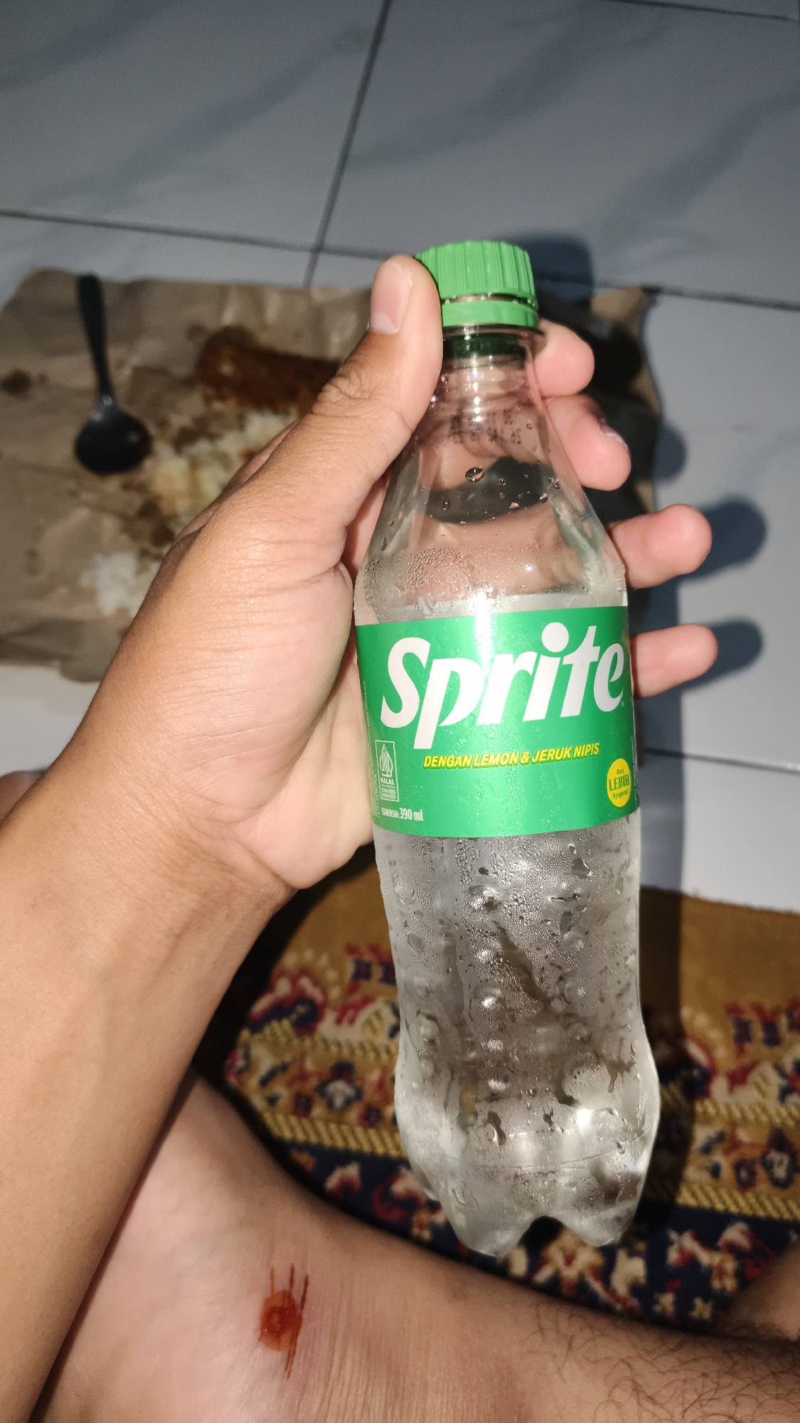 In this image, a person's hand is holding a partially filled, sweating bottle of Sprite. The bottle has a green cap and a label that reads "Sprite" in large white letters. Below that, it says "DENGAN LEMON & JERUK NIPIS" which translates to "WITH LEMON & LIME". There is also a HALAL logo and the volume "390 ml" printed on the label. A small yellow circular sticker with Indonesian text is also visible.
The background shows a tiled floor and a brown paper wrapper with some food and a black spoon on it. The person's foot is also visible in the lower part of the frame, showing a reddish-brown marking or wound on their ankle. The overall setting appears to be indoors, possibly a home, given the tiled floor and the casual presentation of food. The condensation on the bottle suggests the drink is cold. The image is taken in low light conditions, likely in the evening or at night. The presence of food and a beverage implies a meal or snack break. The wound on the ankle is a notable detail. The location context is Barengkrajan, Indonesia.