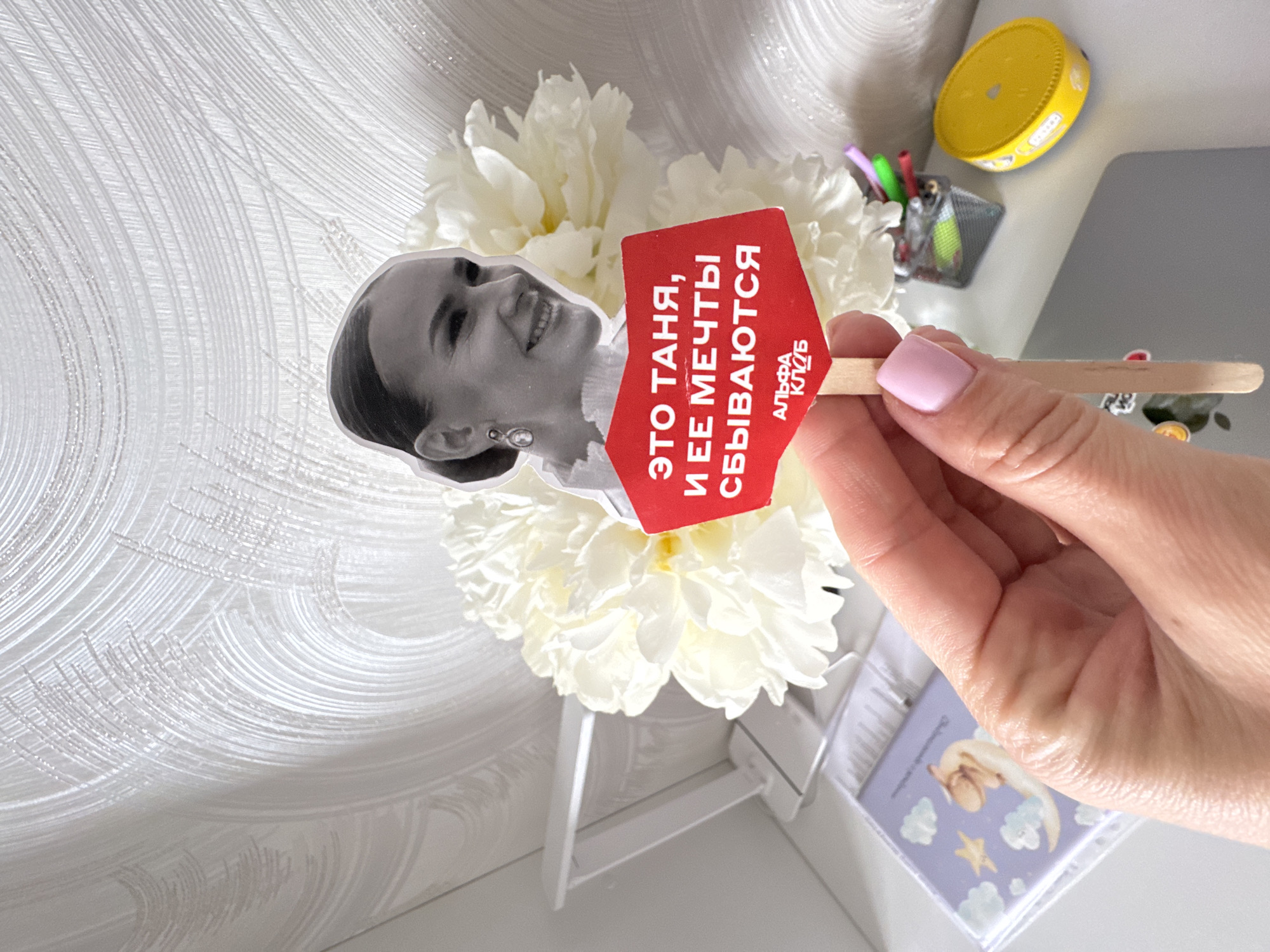 A right hand holds a small, red sign on a stick, displaying a black and white photo of a woman and a slogan in Russian, positioned in front of a bouquet of white flowers. The text on the sign translates to "This is Tanya, and her dreams come true" with "Alpha Club" written beneath. The hand is fair-skinned with light pink nail polish. The bouquet of flowers sits on a small, white stand. In the background, a desk surface is visible with a yellow container, a pen holder with pens, and a laptop. Also visible is a small box with a picture of a baby angel, clouds, and stars on it. The photograph appears to be taken indoors, suggesting a desk or office setting. There is no information available in the image to specify if it is taken in the city of Sochi, Russia.