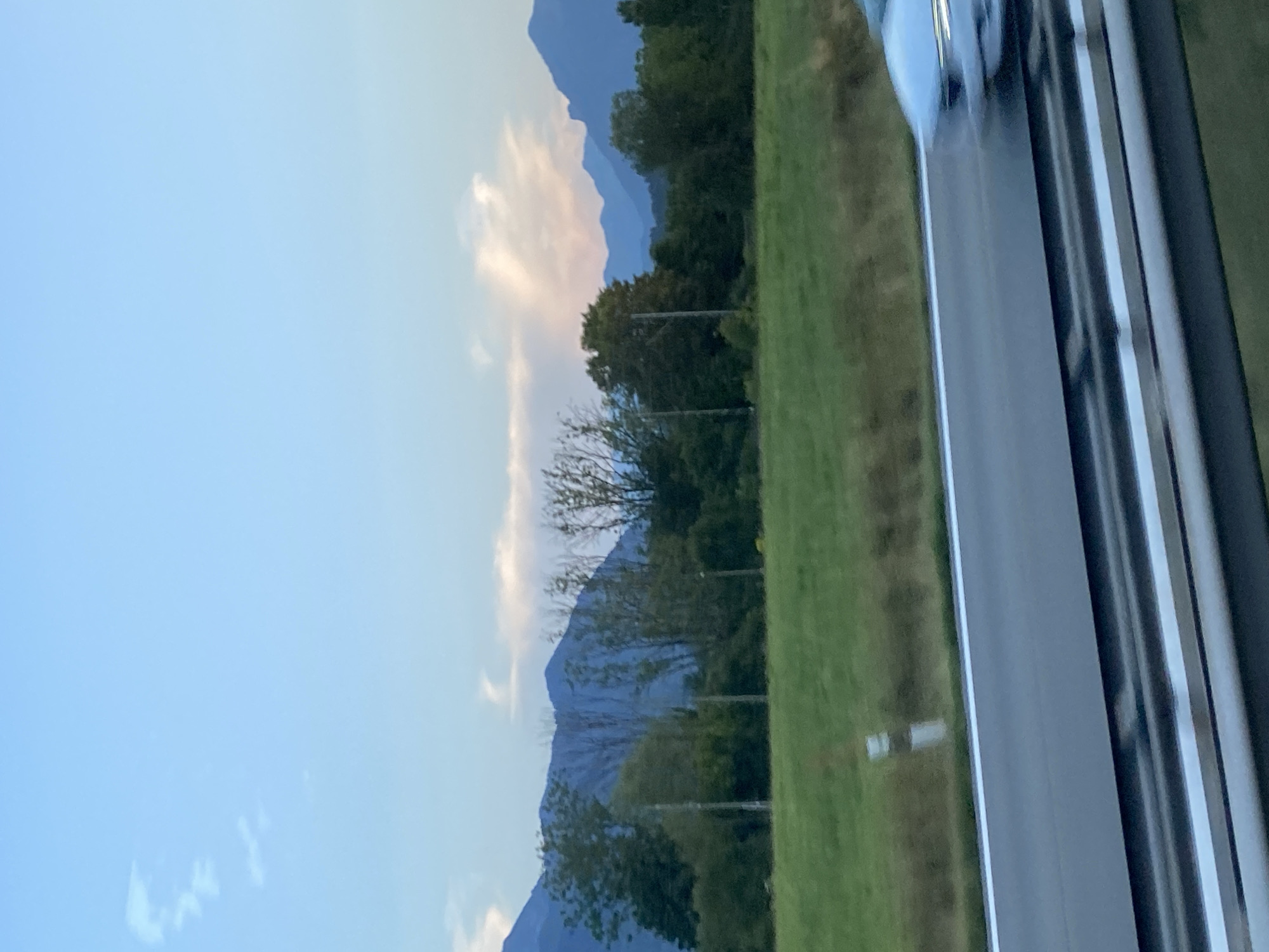 The image depicts a scenic view from a roadside, likely near Grabenstätt, Germany, as indicated by the subtle hint of a mountainous landscape. The foreground is dominated by a road, with a blurred silver car in motion on the right side, suggesting a fast-moving perspective. A metal guardrail separates the road from the viewer's vantage point. Beyond the road, a green, well-maintained field stretches towards a line of trees. In the background, a range of blue-hued mountains forms the horizon, silhouetted against a pale blue sky. Wisps of clouds, tinged with pink and white, are visible near the mountain peaks, hinting at either dawn or dusk. The overall atmosphere is serene and captures a moment of travel through a picturesque rural setting.