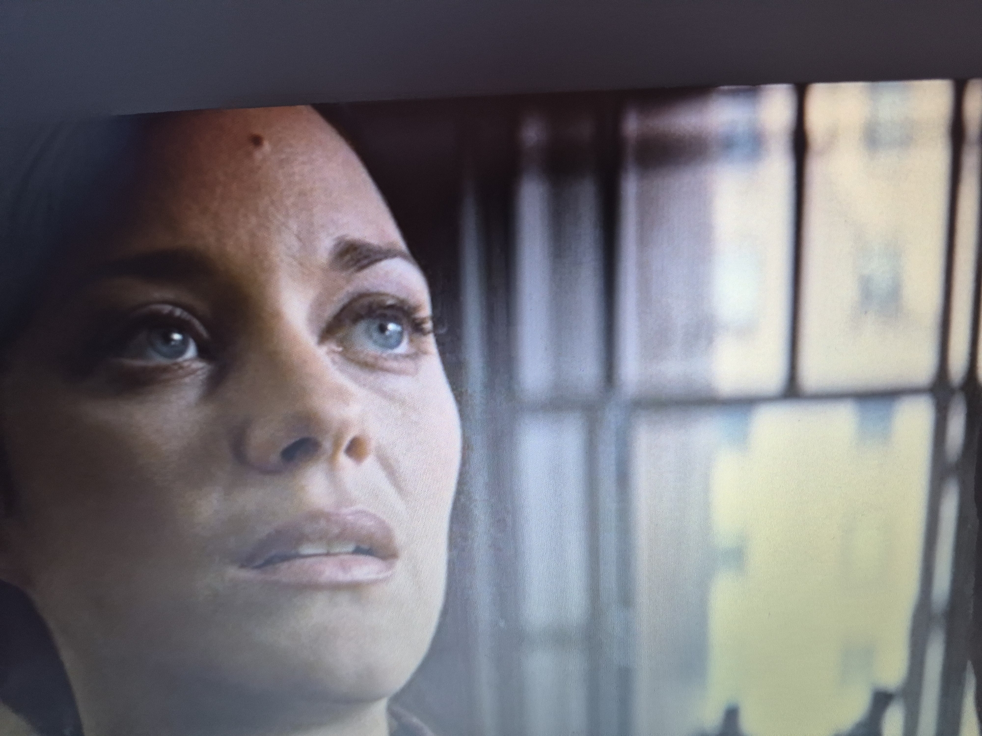 The image is a close-up, slightly out-of-focus shot of a woman's face, specifically her forehead, eye, nose, and mouth. She has light blue eyes and fair skin. A small mole is visible on her forehead. Her expression appears thoughtful or concerned. The background is blurred and appears to be an interior setting with what looks like a window or a grid-like structure, with hints of yellow and muted colors suggesting an urban environment, potentially a building seen through the window. There is no discernible location within the city of Sandwell, United Kingdom.