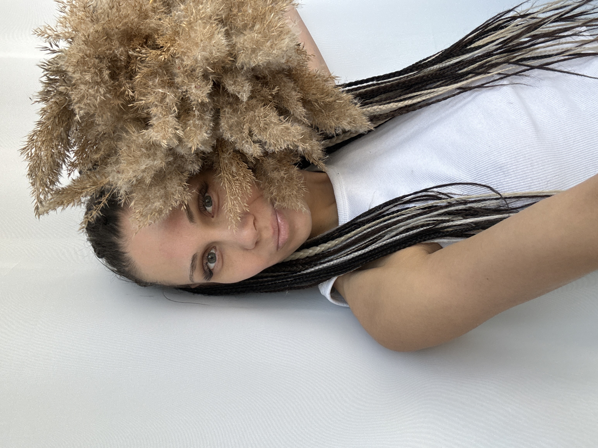 A woman with braided hair lies on a white surface. She is wearing a white shirt and is partially obscured by a large bundle of dried grass. The location is assumed to be Rusanovo, Russia.