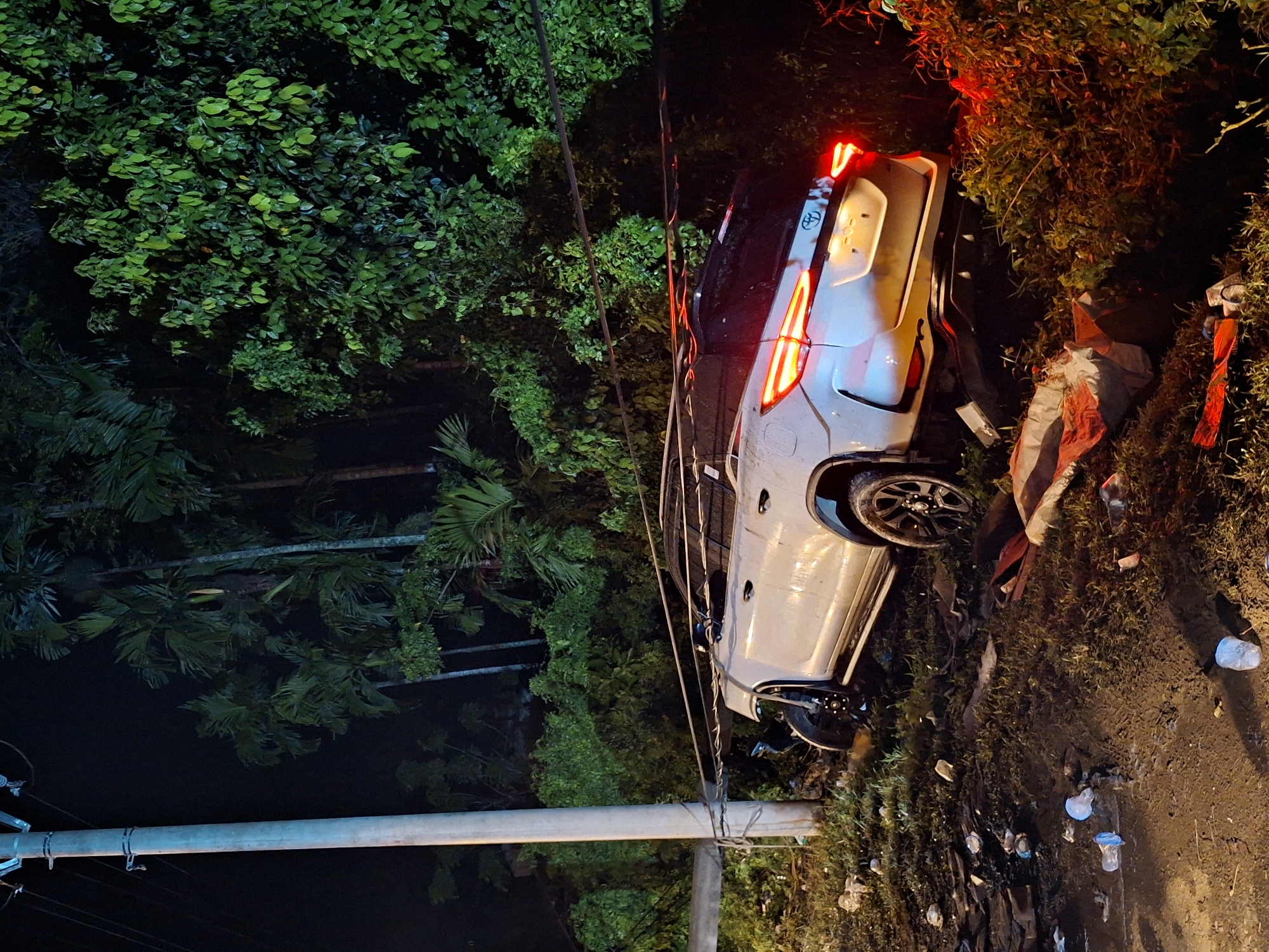 A white SUV lies on its side near a road in Teungoh Baro, Indonesia. The vehicle, identifiable by its taillights and Toyota logo, is surrounded by vegetation and debris. A utility pole with visible wires is positioned near the vehicle. The scene is dark, with light sources coming from the vehicle's lights and potentially street lighting. The general environment suggests a road alongside a vegetation area.