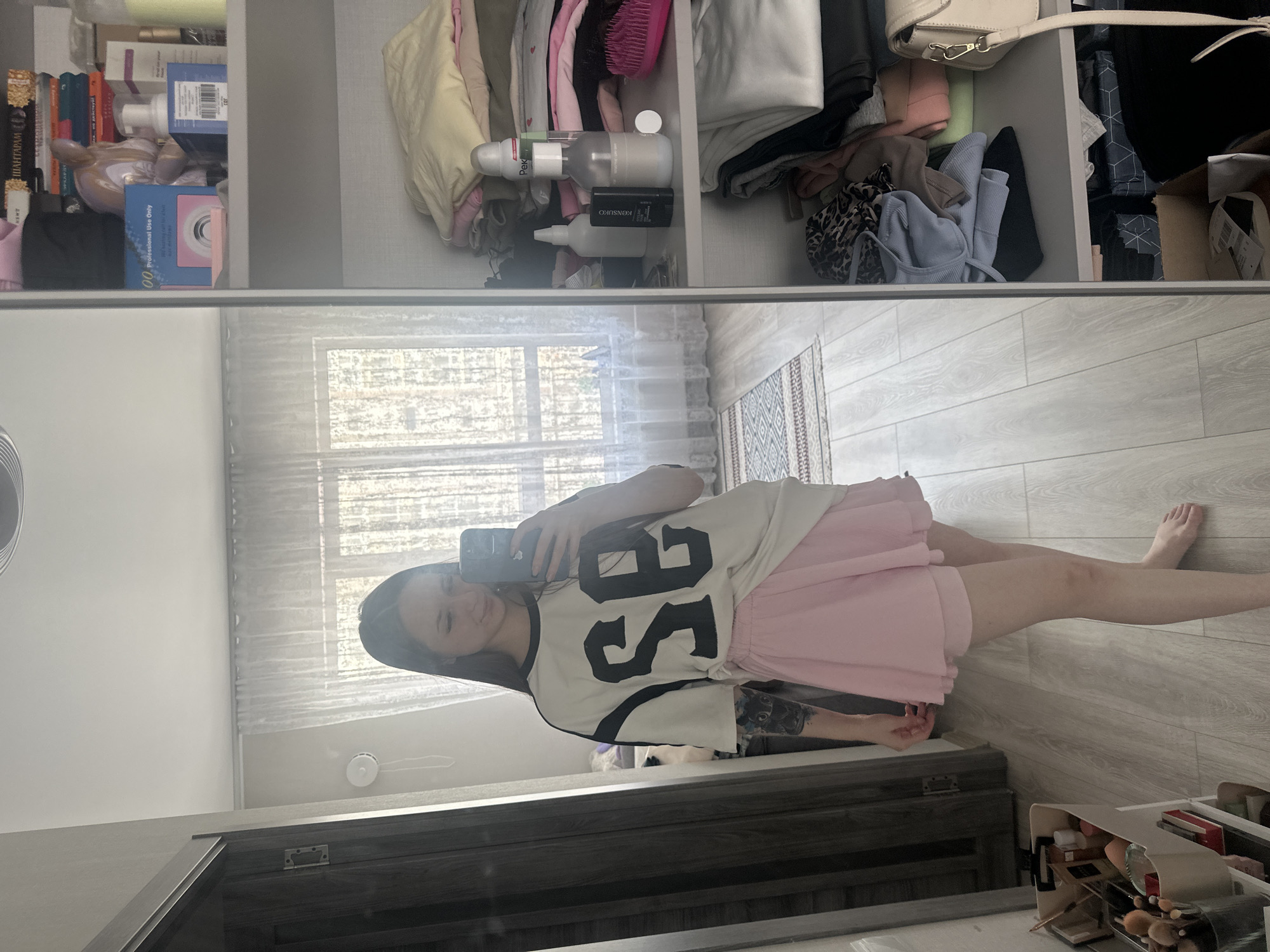 A young woman is captured taking a mirror selfie in what appears to be a bedroom or dressing area. She is reflected from the waist up in a large, possibly full-length, mirror, holding a black smartphone to take the picture. She has long dark hair and a subtle smile. She is wearing a loose-fitting white T-shirt with black sleeves and a large black graphic that appears to be "SE" or "SB" on the front, paired with a light pink skirt. Tattoos are visible on her left arm. Her bare feet and legs are also seen in the reflection.
Above the mirror, a multi-compartment closet or shelving unit is visible, filled with various personal items. The leftmost shelf contains books, including one with the Cyrillic title "ШАНТАРАМ" (Shantaram), alongside several product bottles and a blue box labeled "Professional Use Only." The middle shelf holds more personal care products, including bottles with partially visible text like "PEK" and "KONSURG," a pink hairbrush, and folded light-colored clothing. The rightmost visible shelf contains neatly folded garments in shades of light blue, green, and dark colors, a leopard print item, and a white shoulder bag with a gold clasp.
The room features a light-colored wooden floor and a window covered by sheer white curtains, providing natural light. A white circular smoke detector is visible on the ceiling in the reflection. In the bottom right corner of the actual room, a makeup organizer filled with various brushes and cosmetics is visible on the floor.