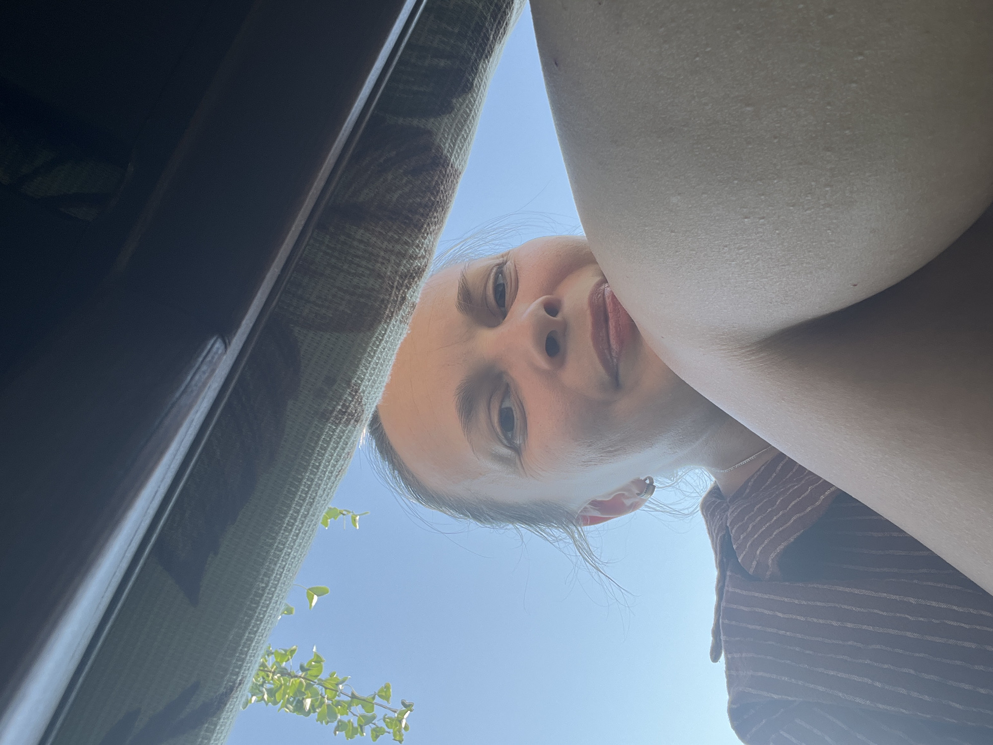 A low-angle, close-up, point-of-view shot shows a woman posing in Lopatino, Russia. The woman has fair skin, brown hair, and brown eyes. She is wearing a brown and white striped shirt. The background is a clear blue sky, with some green leaves hanging from the top left. The woman is leaning forward, with her right arm and shoulder prominent in the foreground. The image is lit by natural light.