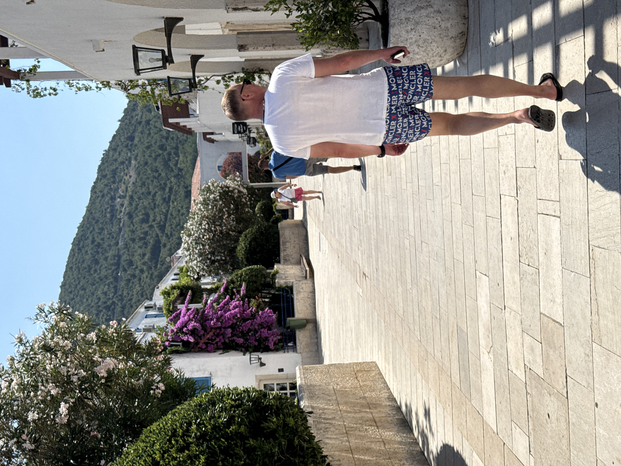 The image shows a man walking on a paved walkway, holding his phone in his right hand. He is wearing a white t-shirt and patterned shorts. In the background, there are buildings and a lush green mountain under a clear blue sky. To the left, there are trees with white and purple flowers. The scene appears to be in a Mediterranean coastal town, with whitewashed buildings and vibrant foliage. The sunlight suggests it is daytime, likely late morning or early afternoon, casting distinct shadows on the ground. The overall atmosphere is relaxed and summery. The shorts the man is wearing have a repeating pattern that appears to be text, possibly a brand name, "MONCLER" is visible.
