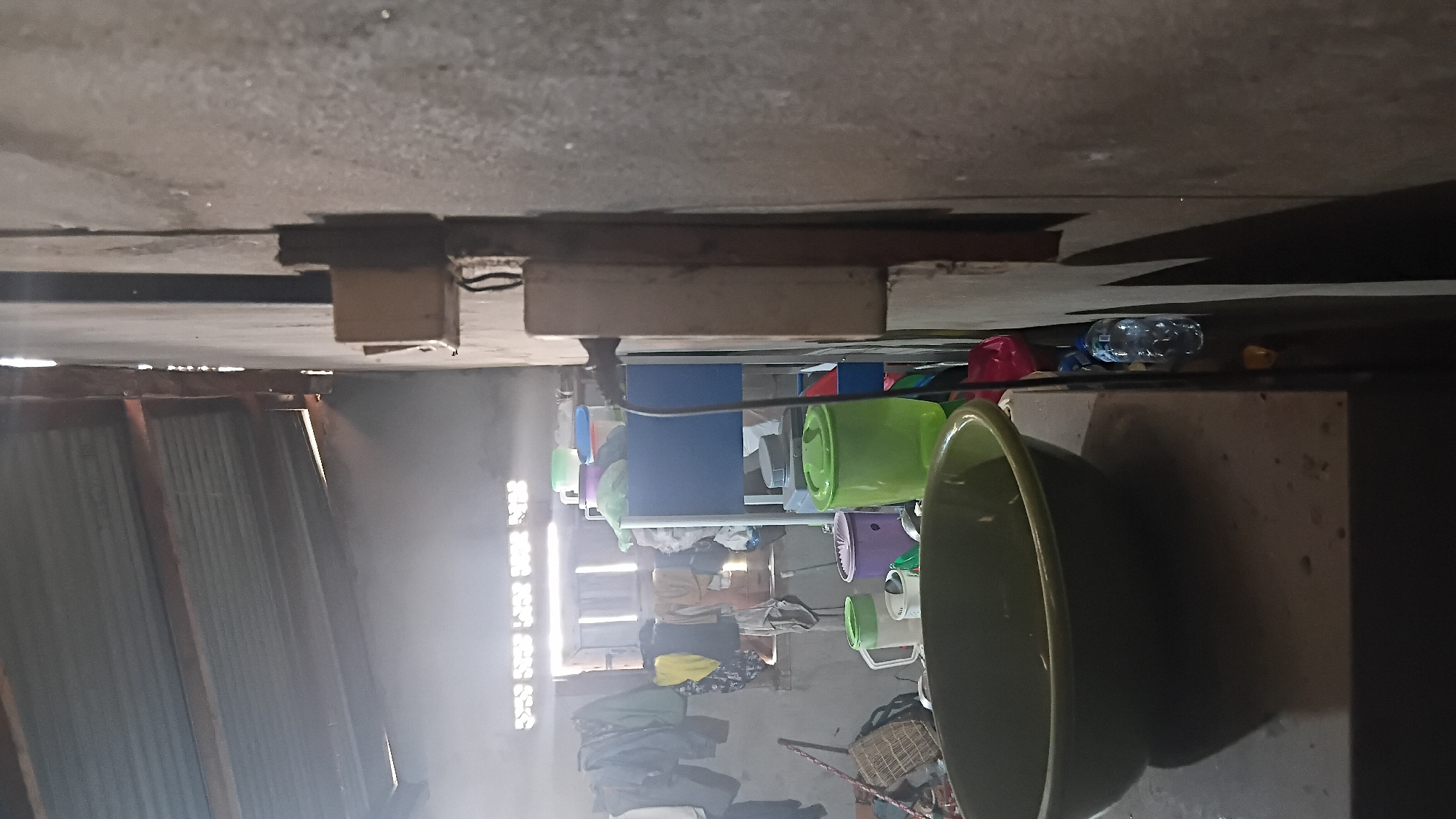 The image depicts the interior of what appears to be a humble residence or workshop in Prabumulih, Indonesia. The scene is cluttered with various domestic items, suggesting a utilitarian living or working space. The focal point in the foreground is a large, dark green basin placed on a countertop. Behind the basin, various containers are visible, including a large green bin, a purple bin, and a white pitcher with a handle. A blue cart with multiple shelves is laden with more containers and what appear to be plastic bags or fabric. Clothes are hanging on a line to the left of the frame, indicating laundry activities. In the background, a window with visible louvers allows light to enter, illuminating dust particles in the air, possibly indicating a smoky or hazy environment. The walls are roughly constructed, with one wall being a textured concrete surface and another appearing to be made of corrugated metal. Electrical components, including a socket and wires, are mounted on the wall to the right. A plastic water bottle is also visible on a surface to the right of the green basin. The overall impression is one of a lived-in, possibly modest, environment in Prabumulih.