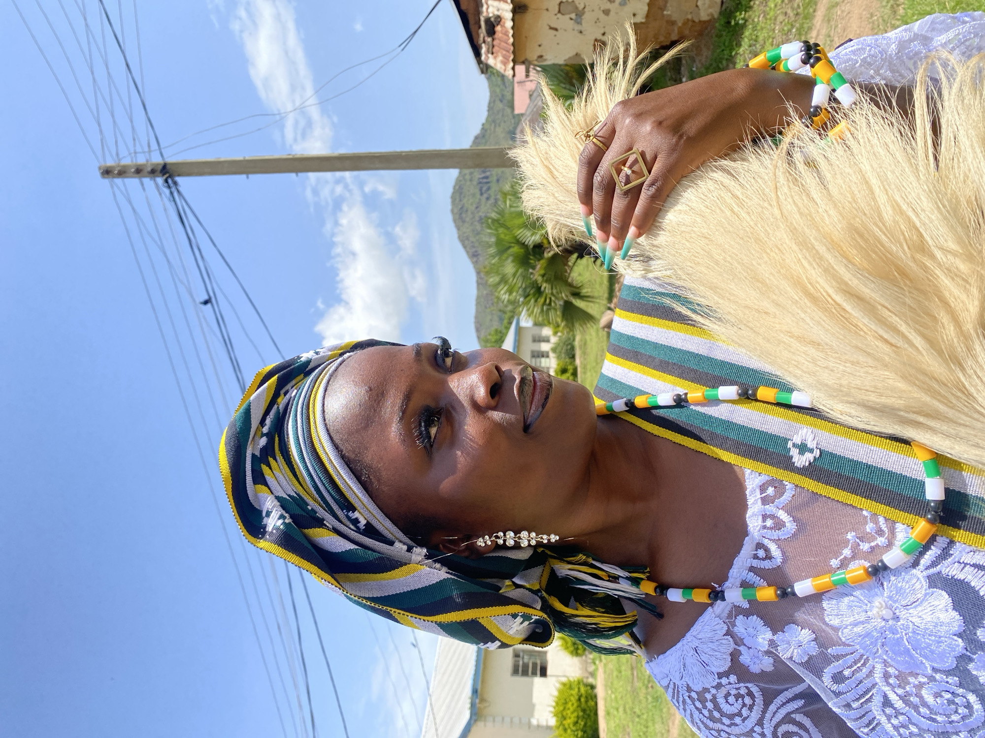 A portrait of a Black woman wearing traditional Nigerian attire, posing outdoors. She has a headscarf wrapped around her head, featuring a geometric pattern of green, yellow, white, and black stripes. Her attire includes a white lace top and a dress with a similar striped pattern. She is adorned with colorful beaded necklaces and bracelets in green, yellow, white, and black beads. A unique geometric ring with a diamond accent is visible on her right hand. She also wears delicate, sparkling earrings. Her fingernails are painted in a gradient of light blue and green. The woman is holding a fluffy, pale yellow fan made of what appears to be hair or fibers.
The background suggests a rural or semi-rural setting in Wang, Nigeria. A utility pole with multiple wires is prominent in the left of the frame, against a bright blue sky with scattered white clouds. Green trees and vegetation are visible, along with a simple building with a corrugated metal roof. The lighting indicates it is daytime, likely sunny. The woman's gaze is directed upwards and to the side, with a neutral to slightly contemplative expression. The overall mood is serene and cultural. No visible text is present in the image.