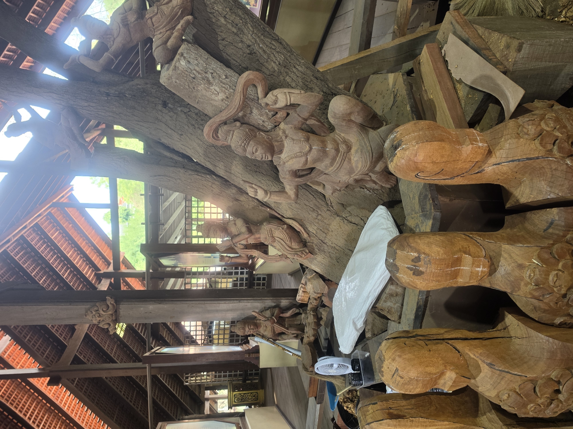 The image depicts an interior space in Pattaya City, Thailand, filled with unfinished wooden sculptures and raw materials. A large, irregular-shaped tree trunk serves as a base for several carved figures, depicting traditional Thai mythological characters, some of which are incomplete. The surroundings are cluttered with various pieces of wood, tools, and a small electric fan. The building structure features a red tile roof and exposed wooden beams, suggesting a workshop or studio environment.