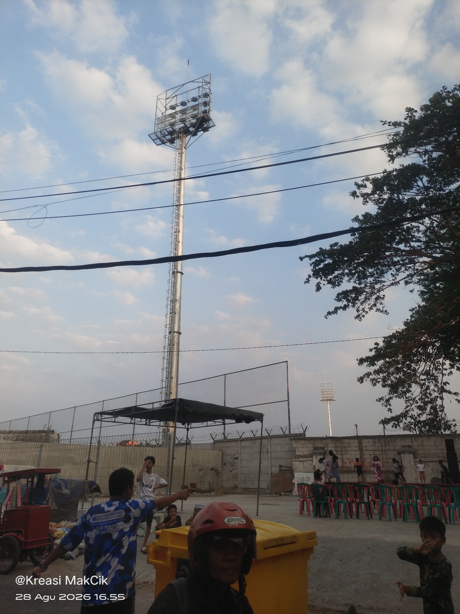 The image captures an outdoor scene at what appears to be a sports field or recreational area in Surabaya, Indonesia. Dominating the background is a tall, metal light tower, likely for illuminating a field at night. The sky is partially cloudy, with a mix of blue and white. In the foreground, there are several people, including children and adults. One adult in a blue and white camouflage shirt is pointing, possibly directing others or indicating something. There are also several rows of chairs set up, some red and some green, facing a wall. A large yellow trash bin is visible in the center foreground. A red cart with a canopy is parked to the left. The overall environment suggests a public gathering or event is taking place or about to take place. The timestamp "@Kreasi MakCik 28 Agu 2026 16.55" is superimposed on the lower left corner of the image.