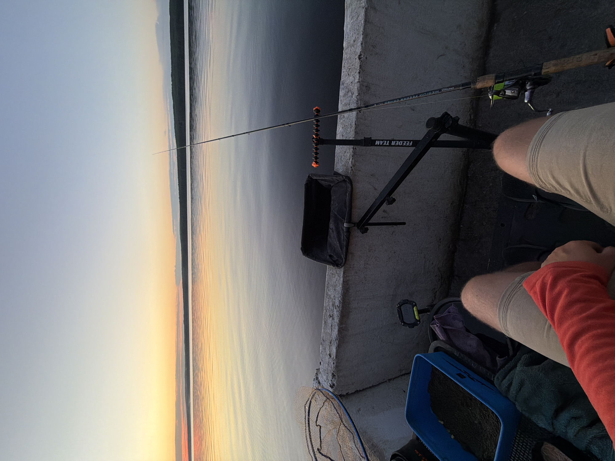 A first-person perspective shot captures a fishing scene at sunset on the Kama River in Chaikovsky, Russia. The foreground features a person's legs clad in beige shorts and a red long-sleeved shirt, seated on a fishing chair. Beside them, a blue plastic container is filled with fishing bait. To the right, a fishing rod is set up on a black feeder arm attached to the chair, its reel visible. A fishing net rests on a concrete barrier to the left. The background is dominated by the tranquil Kama River, reflecting the gradient hues of the sunset sky, with a distant treeline on the horizon. The overall atmosphere is serene and contemplative, indicative of a peaceful fishing outing.