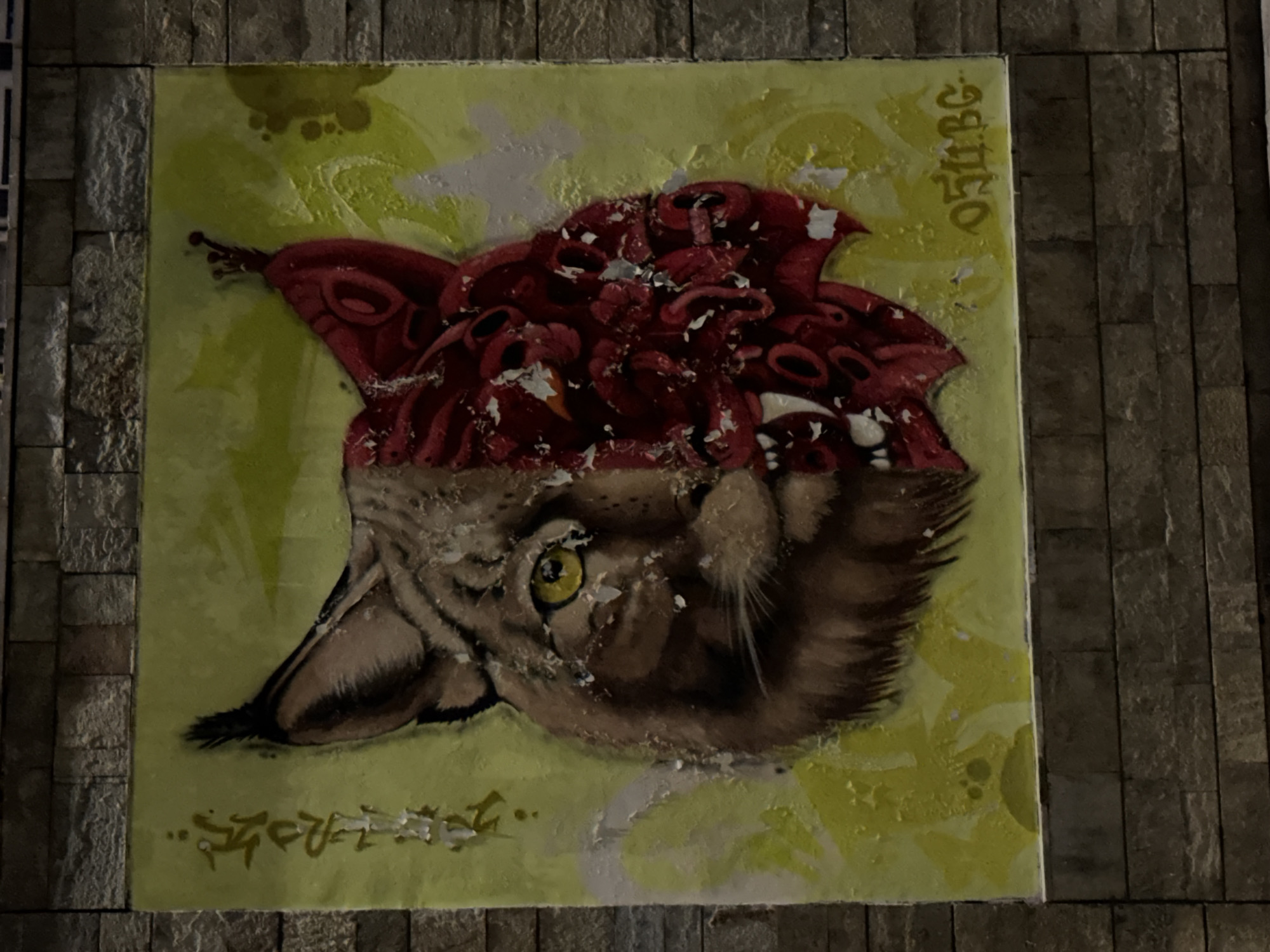 A mural on a brick wall depicts a lynx's head split vertically. The left side is rendered realistically with brown fur and a yellow eye. The right side is abstract and menacing, featuring a red, fanged face with a dark, circular eye and an elaborate, layered structure resembling petals or scales. The background is a textured yellow with green undertones. Faded white spray paint markings are visible on the left side of the mural, and the signature "0511.BG" is spray-painted in yellow on the lower right. The mural is located in Sofia, Bulgaria, on the exterior wall of a building.