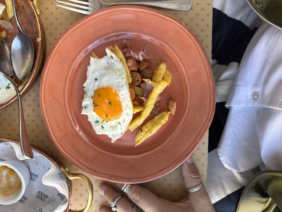 The image displays an overhead view of a prepared meal at Casa Vasco, situated in Aldoar, Foz do Douro e Nevogilde, Portugal. A round, salmon-pink ceramic plate occupies the central foreground, presenting a dish composed of a sunny-side-up fried egg, garnished with chopped chives, layered over golden-brown french fries, sliced sausages, and pieces of cured ham.
To the left of the plate, a person's left hand is visible, adorned with four silver rings: one on the pinky finger, one on the middle finger, and two on the ring finger. Above the hand, a small white ceramic cup containing a foamy coffee beverage rests within a brass-colored metal holder with a handle. This holder is placed on a disposable paper liner featuring the text "CASA VASCO."
On the right side of the main plate, a silver four-tine fork is positioned vertically. In the upper right background, a portion of a second copper-toned metal dish or pan is partially visible, containing remnants of food, including fries and a fried egg, alongside two silver spoons. The table surface is light-colored with a repeating pattern of small reddish-brown star-like designs.
The lower section of the image reveals parts of a person's attire: a white collared shirt with a horizontal navy blue stripe, and a section of a green or khaki-colored bag in the bottom-left corner.