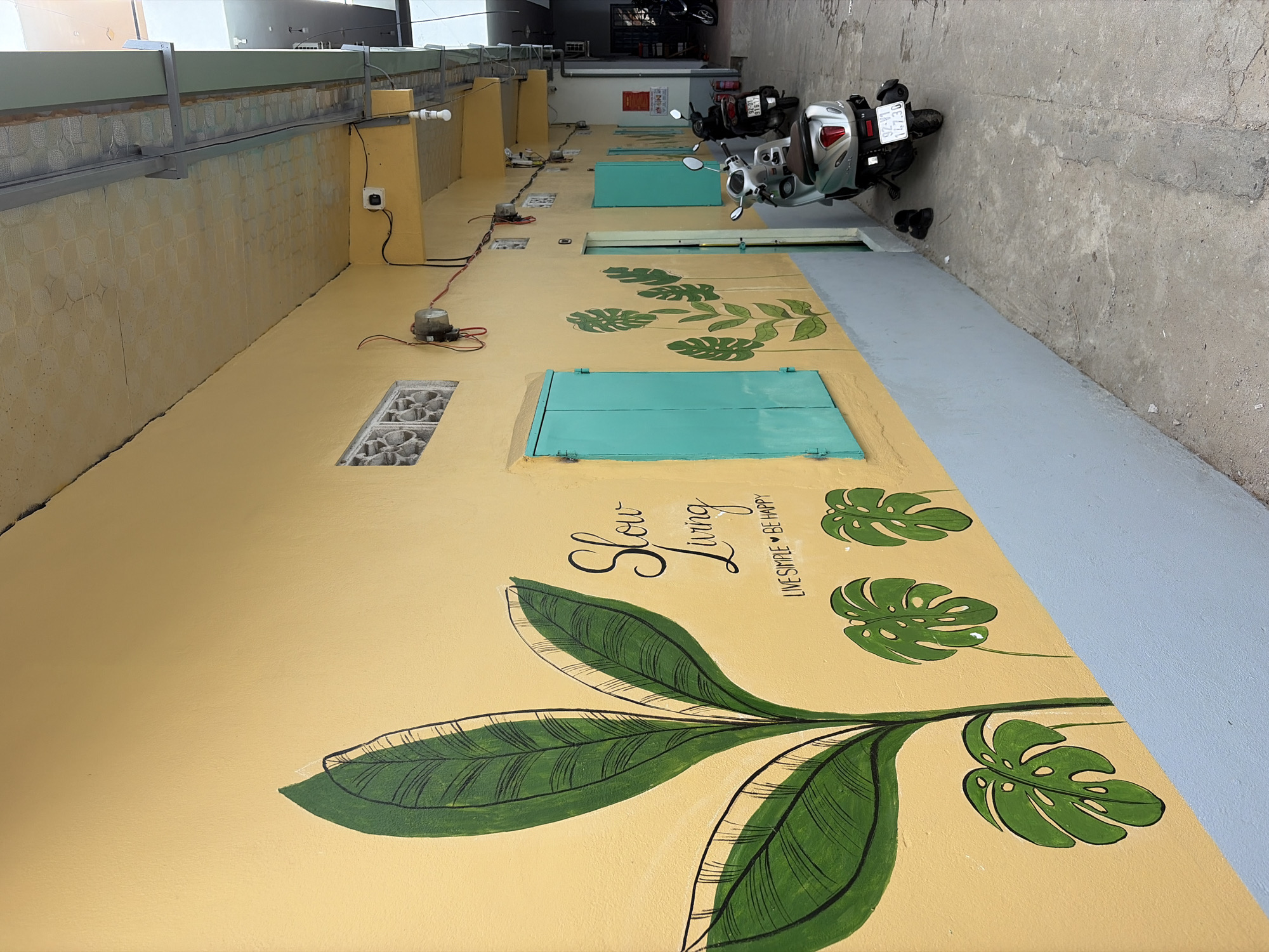 The image shows a narrow outdoor alleyway with a yellow wall adorned with a mural of large green leaves and the text "Slow Living" and "LIVE SIMPLE BE HAPPY". There are two teal-colored doors with shutters on the wall, and a few smaller green leaf drawings. The ground is concrete and a bit cracked.
In the foreground, there is a silver scooter parked on the right side, facing away from the camera. To the left of the scooter, there is a pair of black sandals on the ground. Further down the alley, other parked scooters are visible, some of them partially obscured.
The wall itself has some electrical conduits and fixtures running along it, including what appears to be a security camera. A small barred window is visible near the top of the wall. The ceiling of the alleyway is a light-colored tiled structure with metal framing.
The lighting suggests it is daytime, with a mix of natural light and possibly some artificial light sources from the building or alley.
Based on the style of architecture and the presence of scooters, common in Southeast Asia, and the specific license plate format (e.g., 92-B1 147.30), the location is likely a residential or commercial area in Đà Nẵng, Vietnam. The overall impression is one of a simple, perhaps slightly run-down, urban environment that has been enlivened by decorative art.