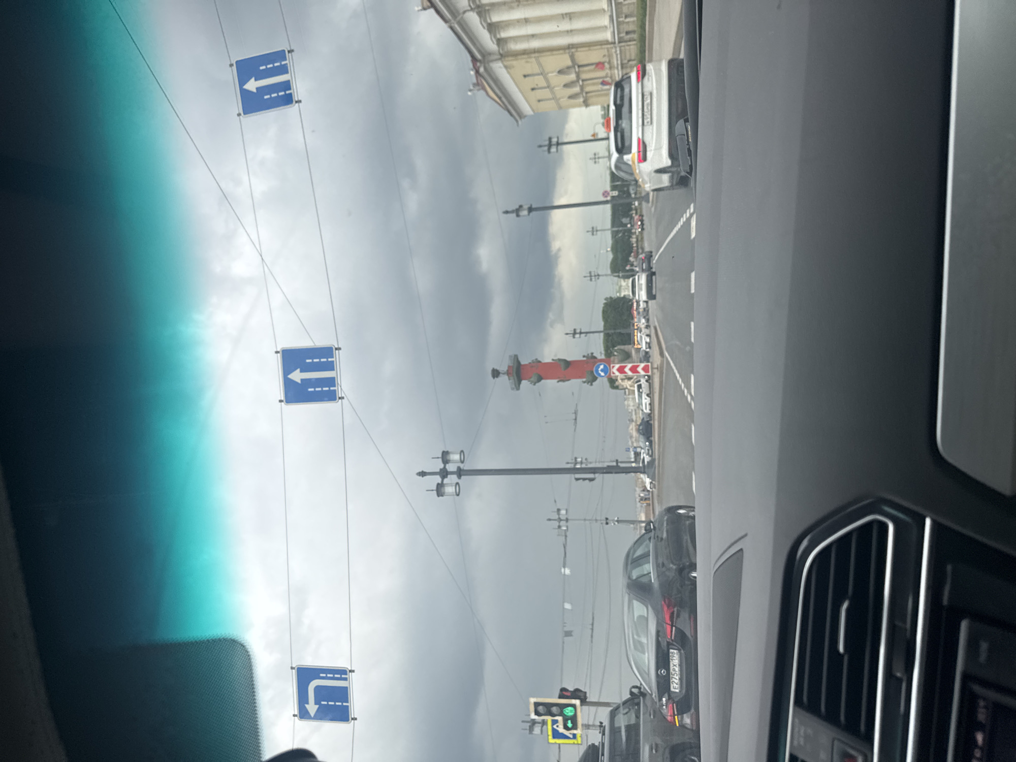 The image is taken from the interior of a vehicle, looking forward through the windshield. It depicts a street scene in Saint Petersburg, Russia, under an overcast sky.
In the foreground, the dashboard of the vehicle is visible. Beyond the windshield, a multi-lane road is seen with several cars driving. Traffic signs are suspended above the road. On the left, a blue square sign with a white arrow pointing left is visible. In the center, a similar sign with a white arrow pointing upwards is present, and another identical sign is on the right.
Further down the road, traffic lights are visible, with the left arrow signal illuminated green. A tall, red column-like monument, likely the Rostral Column, stands prominently in the middle distance. Several lampposts are also scattered along the street.
Buildings with classical architecture, characterized by columns and archways, line the right side of the road. People can be seen walking in the distance near the buildings. The overall environment suggests a city street on a cloudy day.