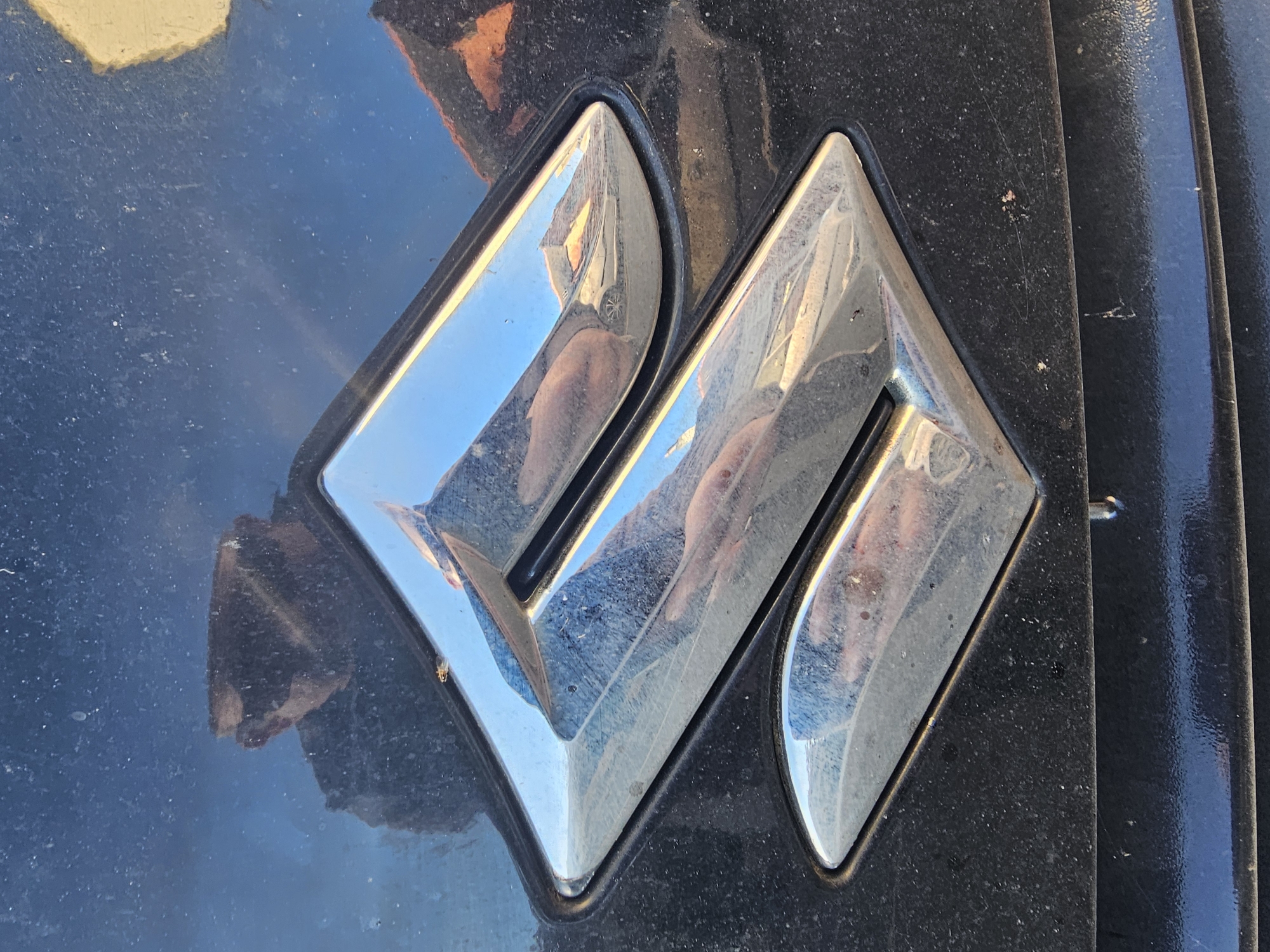 The image provides a close-up view of a chrome vehicle emblem, set against a dark, likely black, and visibly dusty and scratched metallic surface, characteristic of a car's exterior. The emblem is a stylized, angular design, strongly resembling the modern logo for Mahindra, featuring interconnected, diamond-like shapes that form a unique 'M' or 'W'.
The polished chrome surface of the emblem reflects its surroundings vividly. Prominently visible are reflections of a bright blue sky and what appears to be part of a building or another vehicle in the background. Most notably, the distinct reflection of the photographer's arm, hand, and possibly part of their body, holding what seems to be a camera or phone, can be seen across the emblem's surface.
The dark paintwork around the emblem shows a significant layer of dust, small specks of dirt, and several fine scratches, indicating that the vehicle has been exposed to the elements and has not been recently cleaned. The bright reflections suggest the photograph was taken during daytime, under clear or brightly overcast conditions.
The scene captures a static object on a vehicle, with no specific activity taking place other than the act of photography. The wear and tear on the vehicle's surface suggest it is an older model or has seen considerable use. The location context provided is Holywell, United Kingdom, implying this vehicle is situated there. There is no explicit text visible other than the symbolic form of the emblem itself.