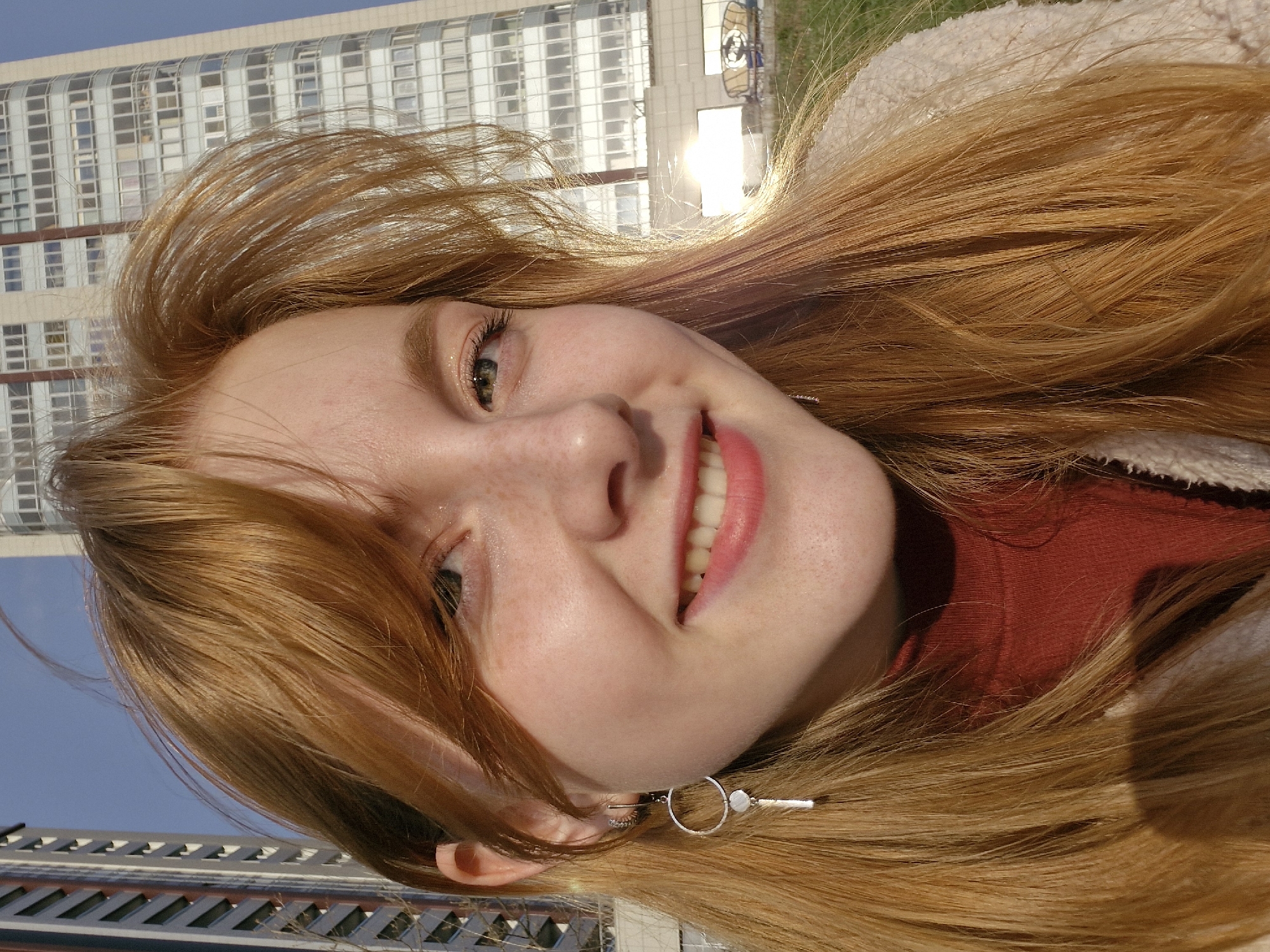 A young woman with long, flowing reddish-blonde hair and freckles is captured in a close-up, outdoor portrait. She is smiling broadly, revealing white teeth, and has green or hazel eyes. She is wearing a red top layered under a cream or light beige fuzzy jacket. A distinctive silver earring with a circular hoop and a dangling bar is visible on her right ear.
The image is taken in bright daylight, indicated by strong sunlight creating a prominent flare in the upper right background. Behind the woman, a modern, multi-story building with numerous windows rises against a clear blue sky, suggesting an urban setting. The sun illuminates parts of the building and casts highlights on the woman's hair. The general location context is Yekaterinburg, Russia. The woman's expression conveys cheerfulness and happiness. There is no visible text in the image.