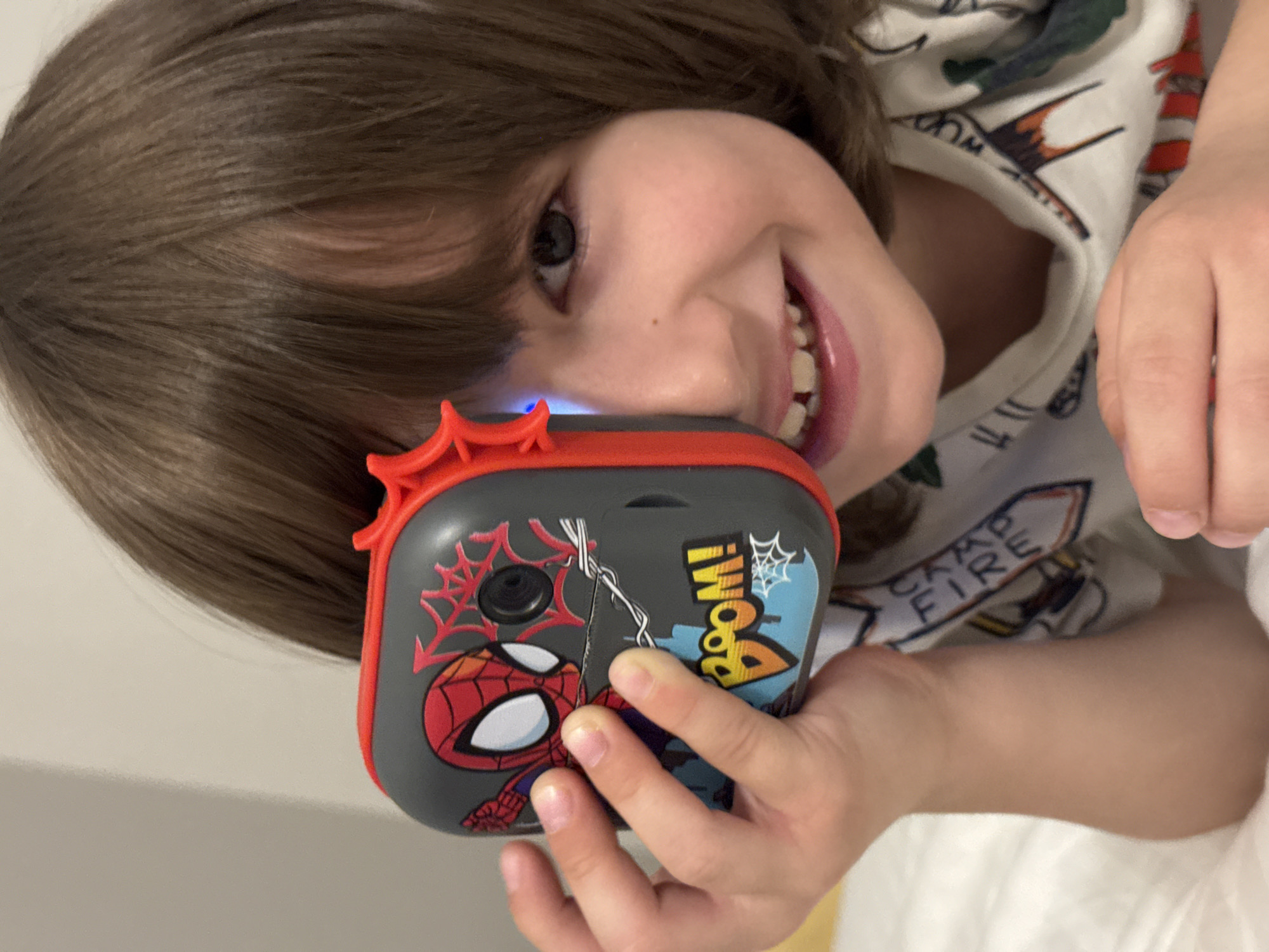 A young child with brown hair and light skin is holding a Spider-Man themed toy camera. The child is smiling with their face tilted towards the camera. The camera is gray with red accents and features an image of Spider-Man on the front, along with the word "BOOM!" in a comic book style. A blue light is visible on top of the camera. The child is wearing a white t-shirt with a print. The background appears to be a white surface, possibly a bed or wall. There is no indication of the location being Chaikovsky, Russia.