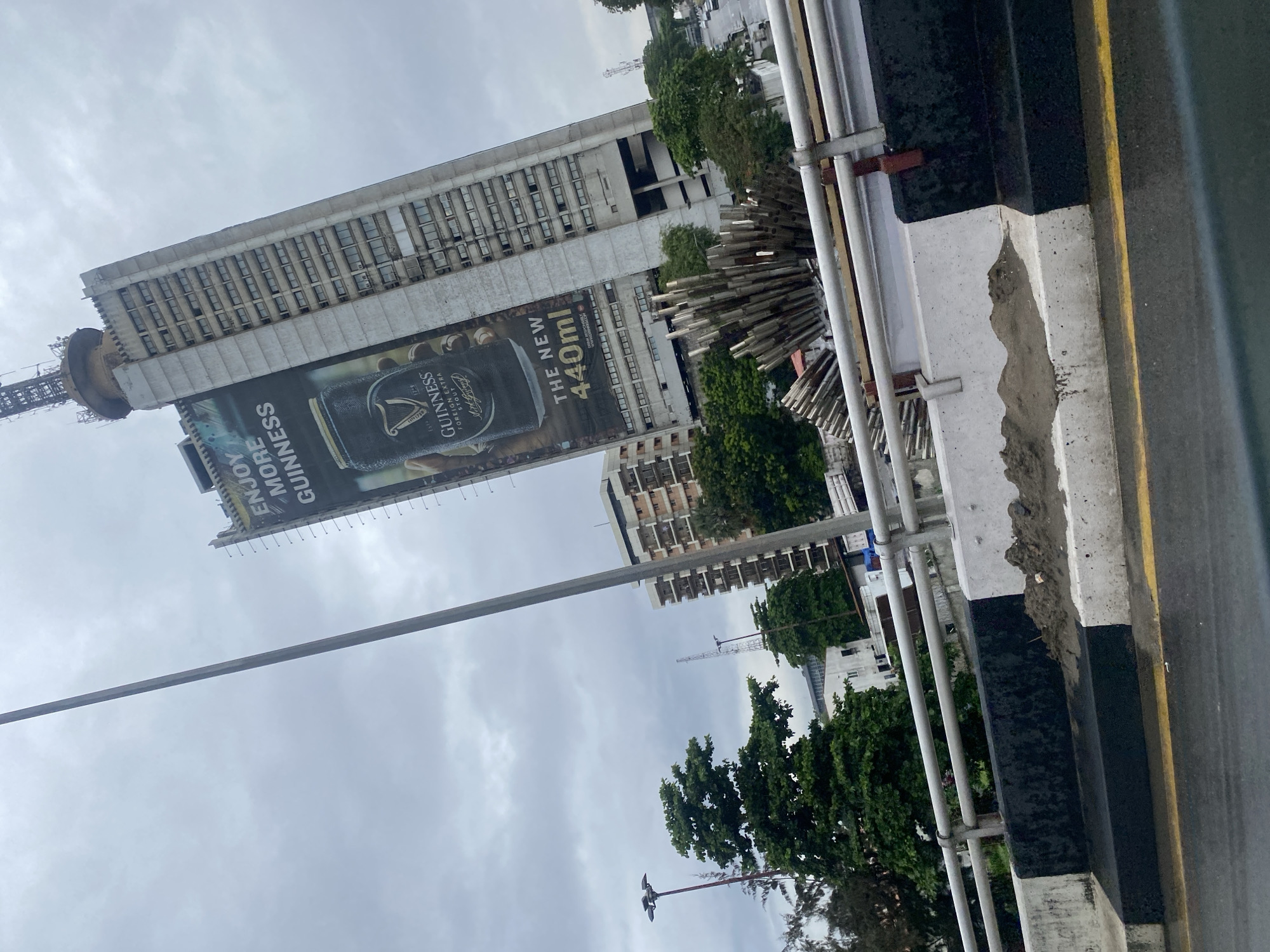 The image shows a wide shot of a city scene on an overcast day. In the background, a tall, imposing skyscraper dominates the skyline, adorned with a large advertisement for Guinness beer. The Guinness ad features a can of the stout and text that reads "ENJOY MORE GUINNESS" and "THE NEW 440ml". To the left of the skyscraper, other buildings and trees are visible. In the foreground, a concrete barrier with black and white markings runs across the frame, separating a road from what appears to be a pedestrian walkway or embankment. The road surface is wet, suggesting recent rain. The scene is located in Victoria Island, Nigeria. There are no people visible in the image, and the focus is on the urban landscape and the advertisement.