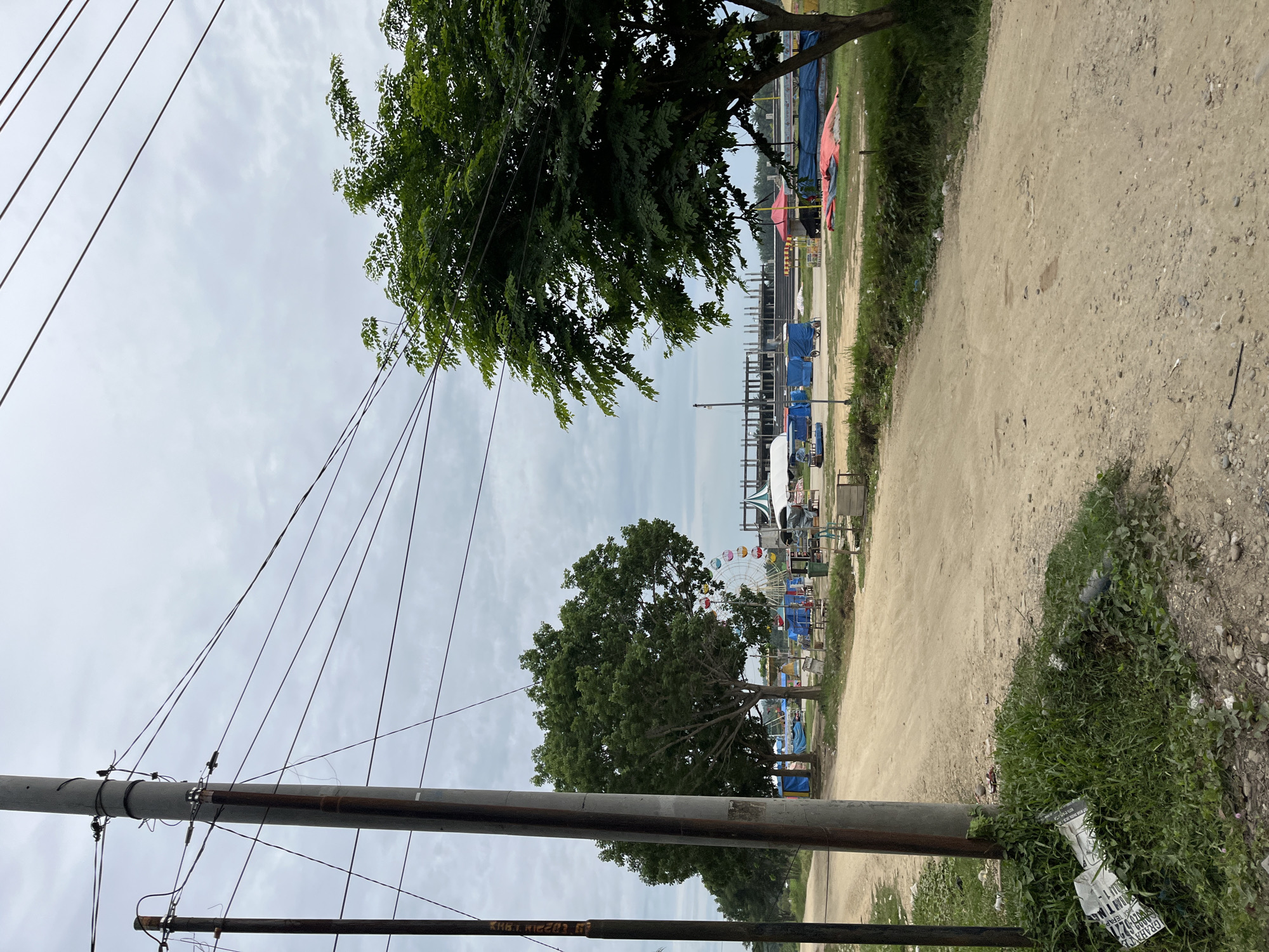 This image captures a street scene in Bundar, Indonesia, under an overcast sky. A dirt road dominates the foreground, with a prominent utility pole on the left side of the frame adorned with numerous electrical wires that crisscross the sky. To the right of the road, a lush green tree with dense foliage extends into the frame.
In the middle ground, a Ferris wheel with colorful pods stands partially obscured by trees. Beyond the Ferris wheel, several temporary structures covered in blue tarps are visible, suggesting a market or a gathering. Some small carts or stalls are also present. In the background, the skeletal framework of a large building under construction, possibly a stadium or industrial facility, is discernible against the cloudy sky. The ground appears to be a mixture of dirt and sparse grass, with some scattered debris near the utility pole. The overall atmosphere suggests a developing area, possibly on the outskirts of a town or city.