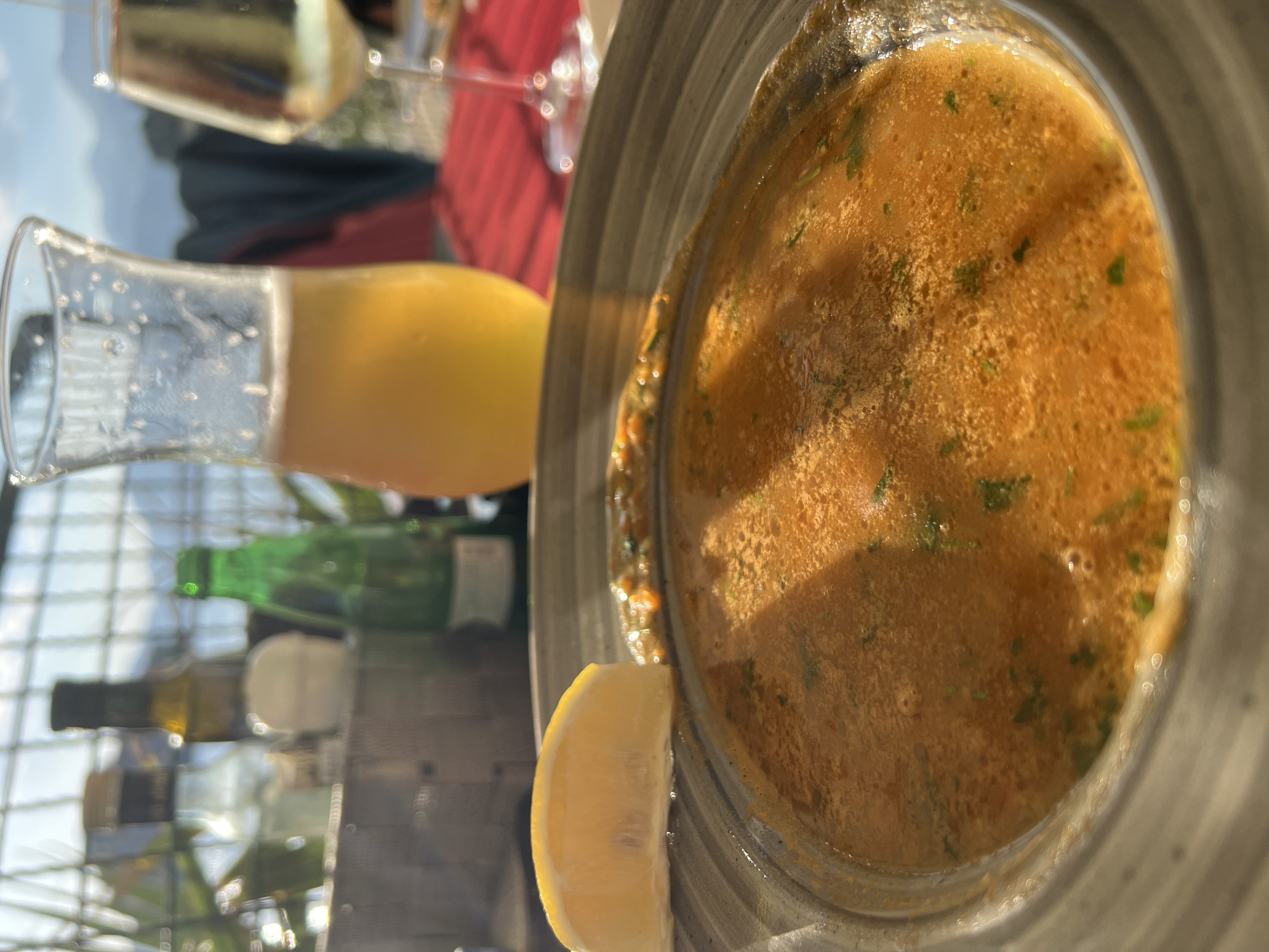 In Sveti Stefan, Montenegro, a close-up shot focuses on a bowl of vibrant orange soup garnished with a lemon wedge, suggesting a meal on a terrace. The background is softly blurred, revealing a table setting with a glass of pale yellow liquid, likely beer, and a wine glass with a white beverage. Behind the table, a metal railing encloses the outdoor space, with glimpses of greenery, bottles, and distant mountains under a bright, hazy sky. The lighting indicates it's daytime, possibly late afternoon, casting shadows across the soup.