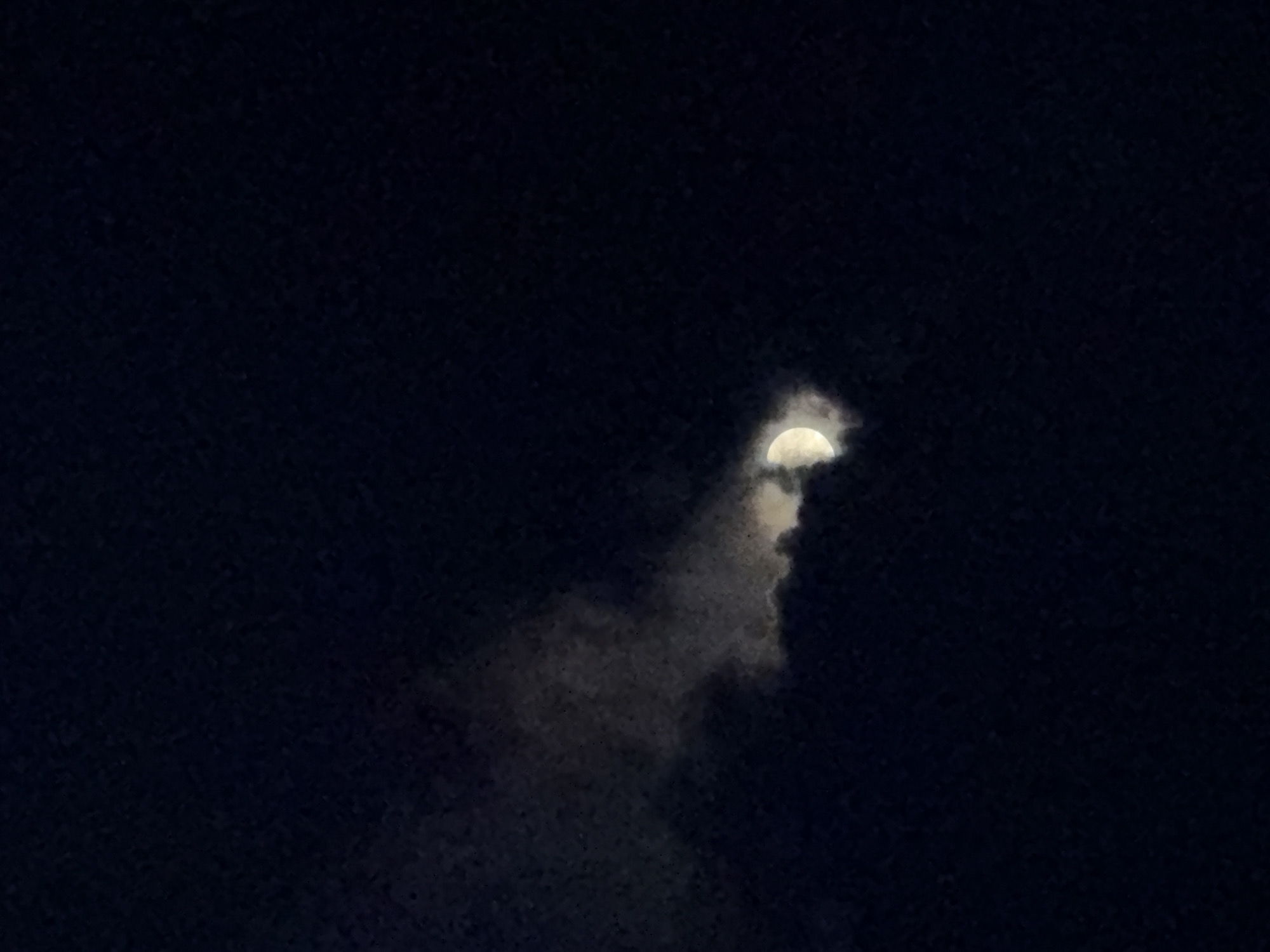 A crescent moon is partially obscured by dark clouds against a deep blue night sky. The clouds are wispy and appear to be illuminated by the moon's light. The image is a close-up shot, focusing on the moon and clouds, with the surrounding sky being uniformly dark. There is no discernible location in Garmisch-Partenkirchen, Germany, as the image only depicts celestial elements. There are no people, subjects, objects, actions, or interactions visible in the image. The environment is solely the night sky with cloud cover.