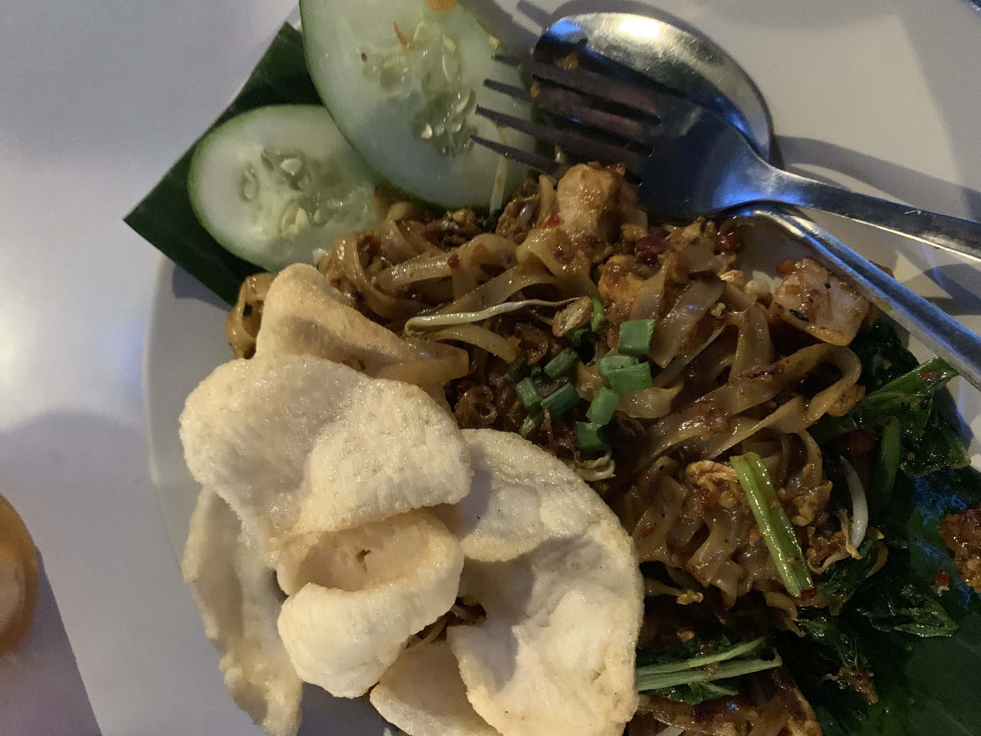 A plate of Pad See Ew with shrimp chips and cucumber slices on a banana leaf is presented in Tamanbaru, Indonesia. A spoon and fork rest on the right side of the plate. Green onions and bean sprouts are also visible in the dish.