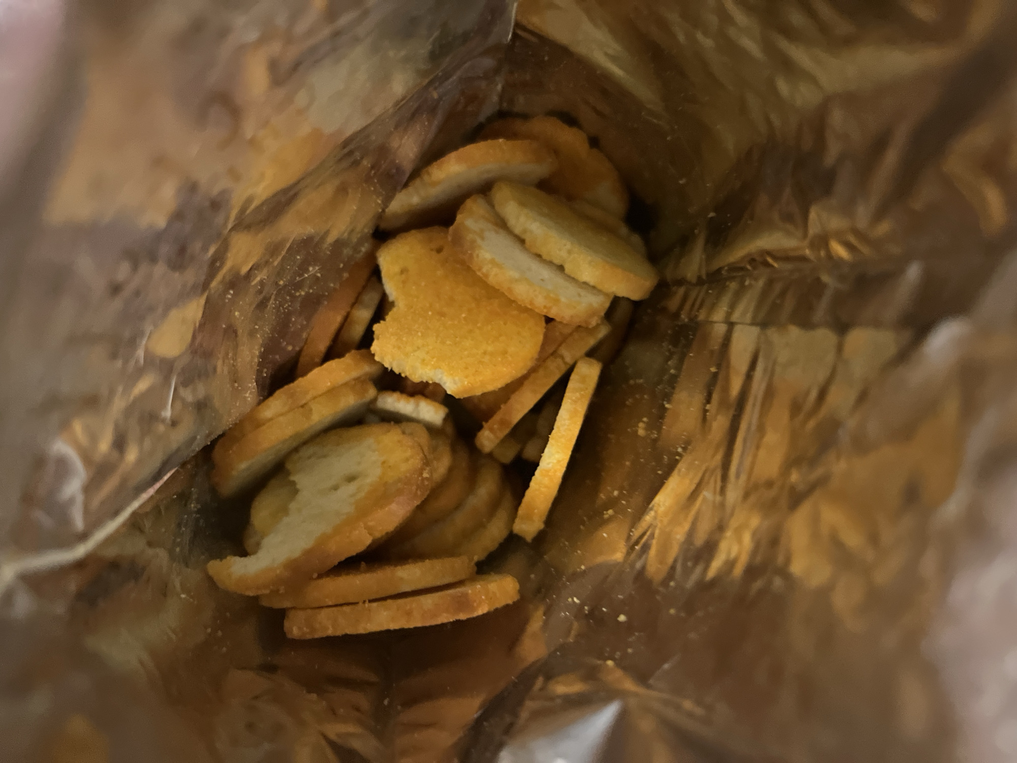 The image is an overhead, close-up shot looking down into a shiny, golden bag, likely made of foil, filled with sliced bread. The bread slices are arranged in a jumbled pile, with some overlapping and others lying flat. The slices have a pale interior and a golden-brown crust, with some appearing to have a seasoning or coating of orange powder. The bag is slightly open, and the interior is reflective, showing highlights and shadows. There are no people or animals present. The scene is indoors, and there are no discernible city landmarks or environmental details visible that would place it specifically in Vratsa, Bulgaria. The focus is solely on the contents of the bag.