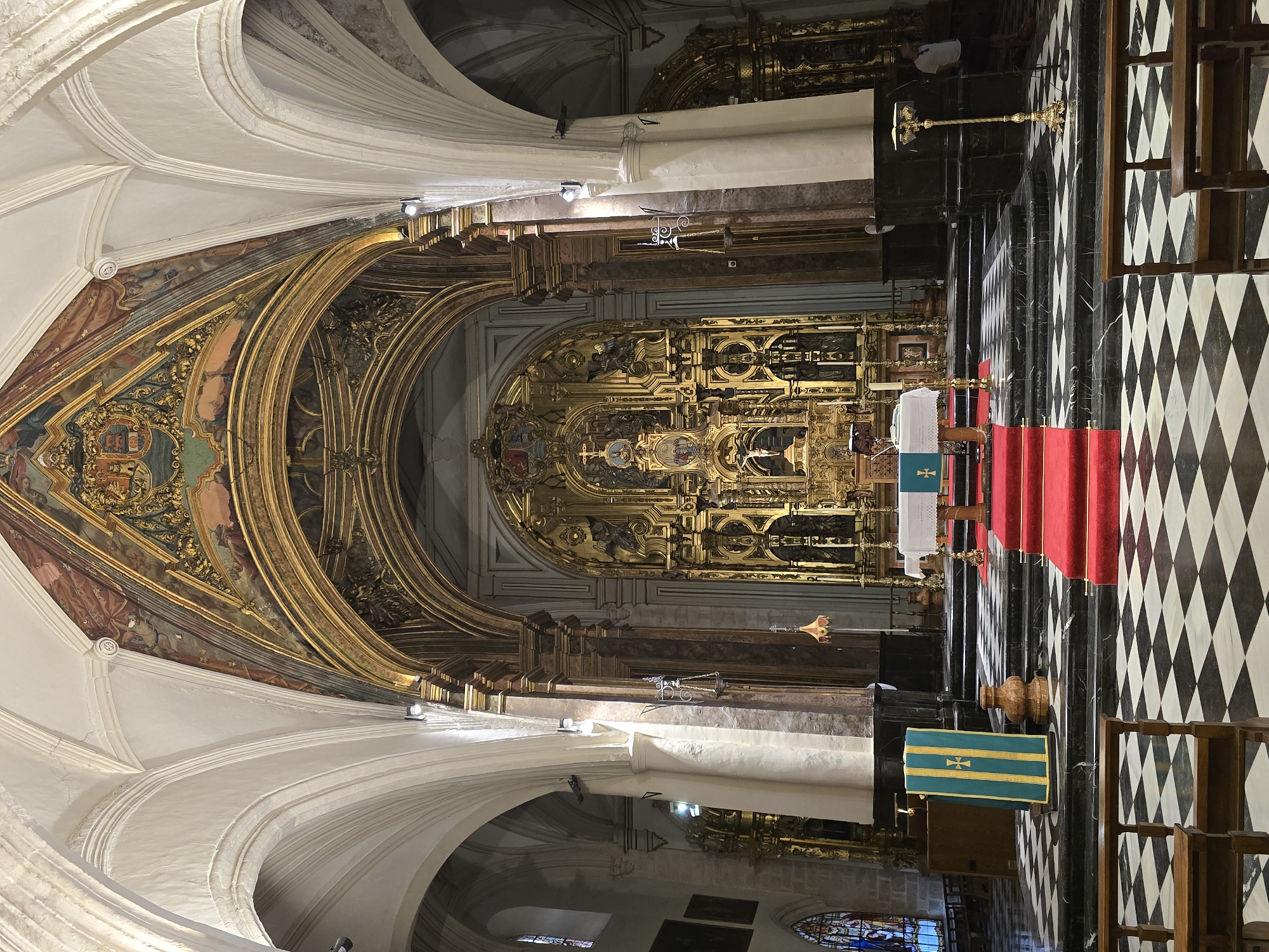 This is an interior shot of a grand, ornate church, likely the Cathedral of Jaén, Spain. The perspective is looking towards the altar, which is a large, gilded retablo featuring numerous sculptures and architectural details. A red carpet runs up a set of steps leading to a white-covered altar table. The church interior is characterized by high, vaulted ceilings with painted decorations, marble columns, and a black and white checkered floor. Benches for congregants are visible in the foreground. A person in a white shirt and dark pants stands to the right, near a golden lamp stand. Another person is partially visible behind the altar. The overall impression is one of opulence and religious solemnity.
