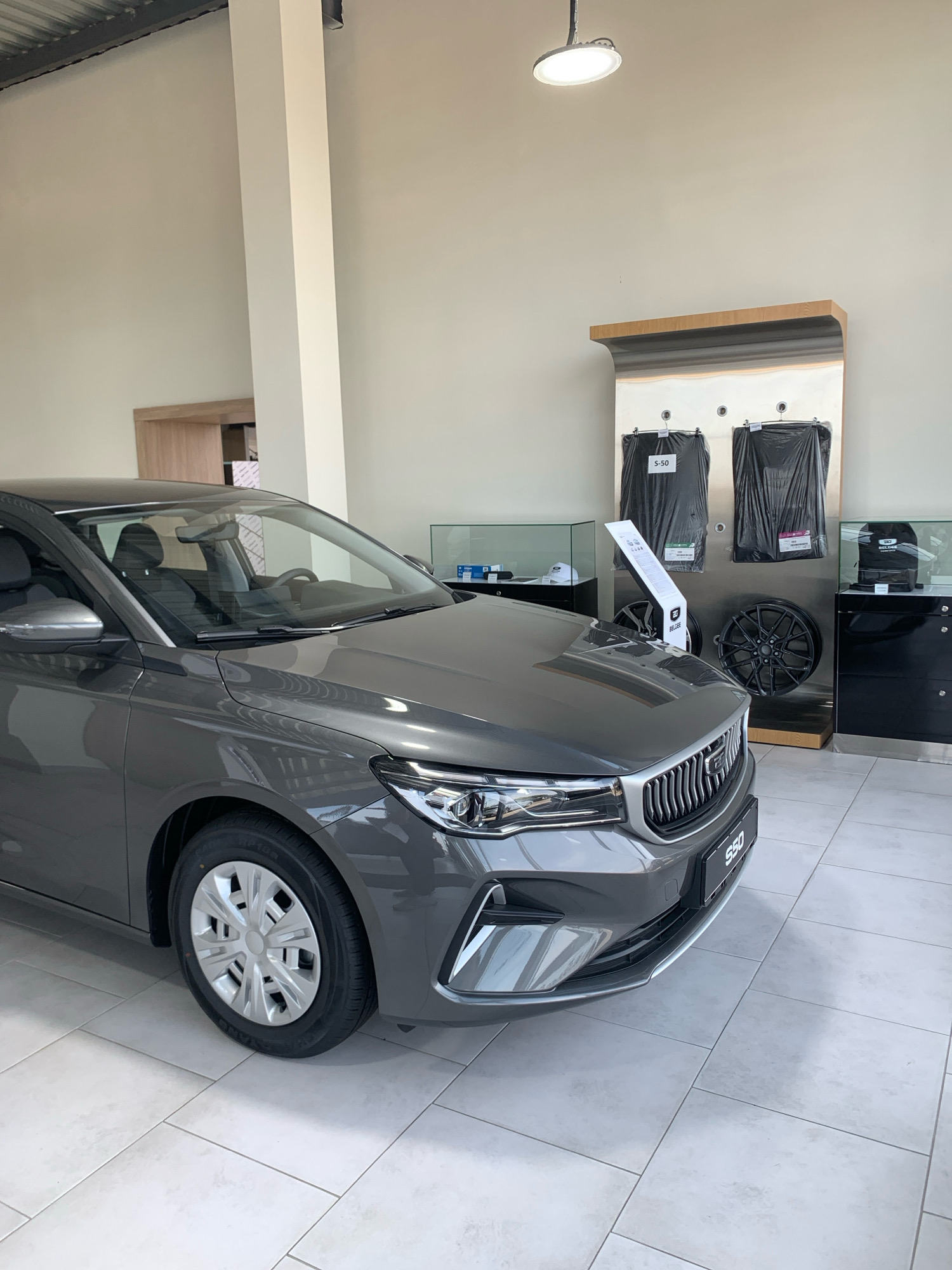 A grey car is parked in a showroom. The car is in the foreground, taking up the left side of the image. It is a sedan with its front driver's side tire visible. The car's headlights and grill are also visible. The interior of the car is dark, with the driver's seat and steering wheel visible through the windshield.
Behind the car, in the background, are several display stands. One stand has two black car floor mats hanging from it, labeled "S-50". Another display shows two car rims. To the right of the car, there is a glass display case with a black backpack inside. The floor of the showroom is tiled in a light color. The walls are painted a neutral color. A white pillar is visible to the left of the car, and a door is visible in the far back. A ceiling light fixture is also visible in the upper right corner. The scene appears to be an automotive dealership. The location is Чыжэвіцкі сельскі Савет, Belarus.