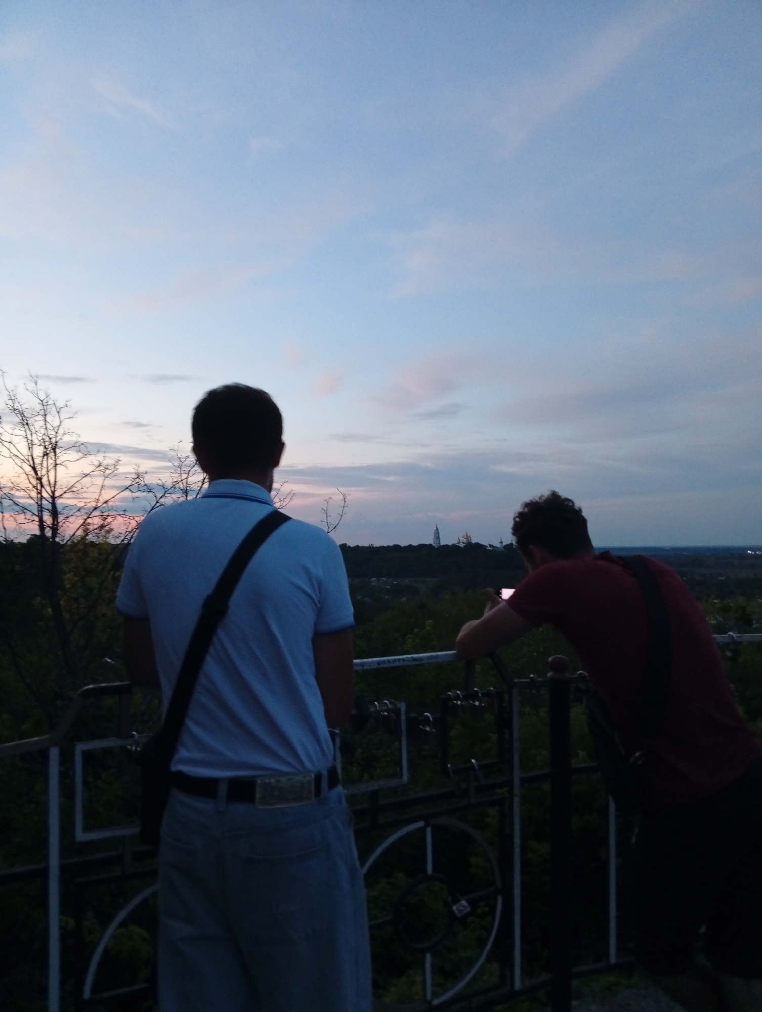 This image shows two people standing on an elevated viewpoint overlooking a landscape at dusk. The sky is a soft gradient of blue, with hints of pink and orange clouds, indicating either sunrise or sunset. The foreground features a dark railing, suggesting a balcony or observation deck.
On the left, a person wearing a light blue polo shirt and light-colored pants is seen from behind, with a dark strap across their chest. They are looking out at the view. On the right, another person in a dark red t-shirt is also viewed from behind, leaning on the railing and holding a phone, appearing to take a picture or record a video.
In the distance, a town or city is visible with trees dotting the landscape. A prominent white structure, possibly a spire or monument, is discernable against the fading light. The overall mood is tranquil and contemplative, with the subjects absorbed in the scenery. The time of day is clearly evening or early morning due to the sky's coloring and the low light. The presence of a railing and the elevated viewpoint suggests a public space designed for sightseeing. No visible text is present in the image.