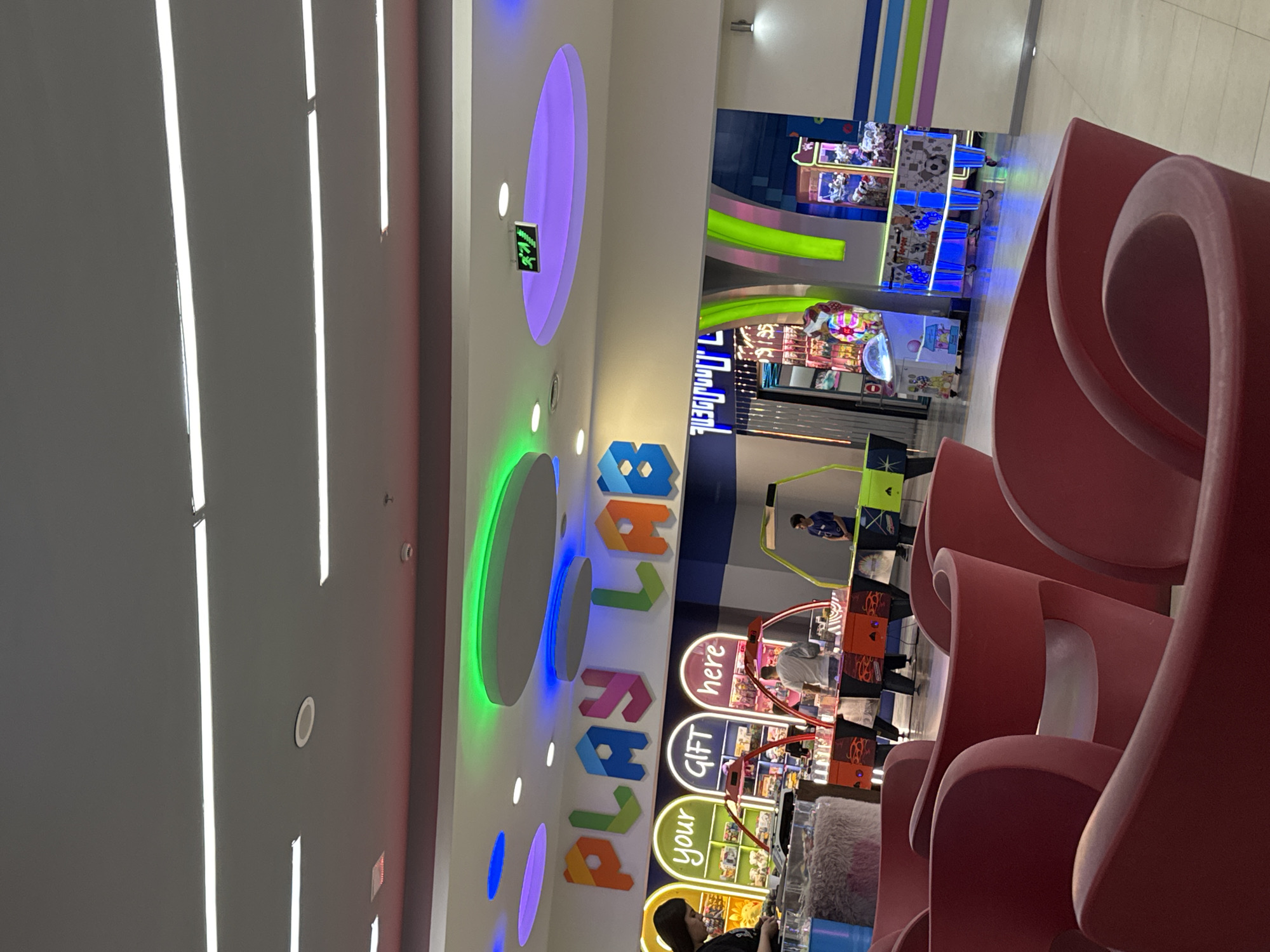 This image captures a vibrant and modern indoor entertainment center, likely an arcade or a play area, situated in Yerevan, Armenia. The scene is brightly lit with a combination of white strip lights on the ceiling and colorful LED lighting, including glowing circular fixtures in green, blue, and purple, contributing to a lively atmosphere.
Several people are present, engaging with various arcade machines and prize redemption games. In the foreground on the right, large, abstractly shaped red seating, possibly made of plastic or fiberglass, curves into the frame, providing a striking visual element. Further into the background, individuals can be seen operating claw machines or similar games, with one person in a blue shirt visible near a game, and another in a light-colored top engaged with a machine that has "here" written on it. A person with dark hair is also visible towards the bottom right, next to what appears to be a fluffy cushion or toy display.
The walls feature colorful paneling and a prominent, multi-colored sign spelling out "PLAYLAB" in large, block letters. Below this, there are three distinct signs above redemption games that collectively read "your GIFT here," suggesting a prize counter or reward system. Another sign in the distance appears to say "DREAM DEAL," although it is partially obscured. The general activity indicates a leisurely setting where people are enjoying games. The time of day is not discernible due to the indoor lighting, but the ambiance is cheerful and inviting.