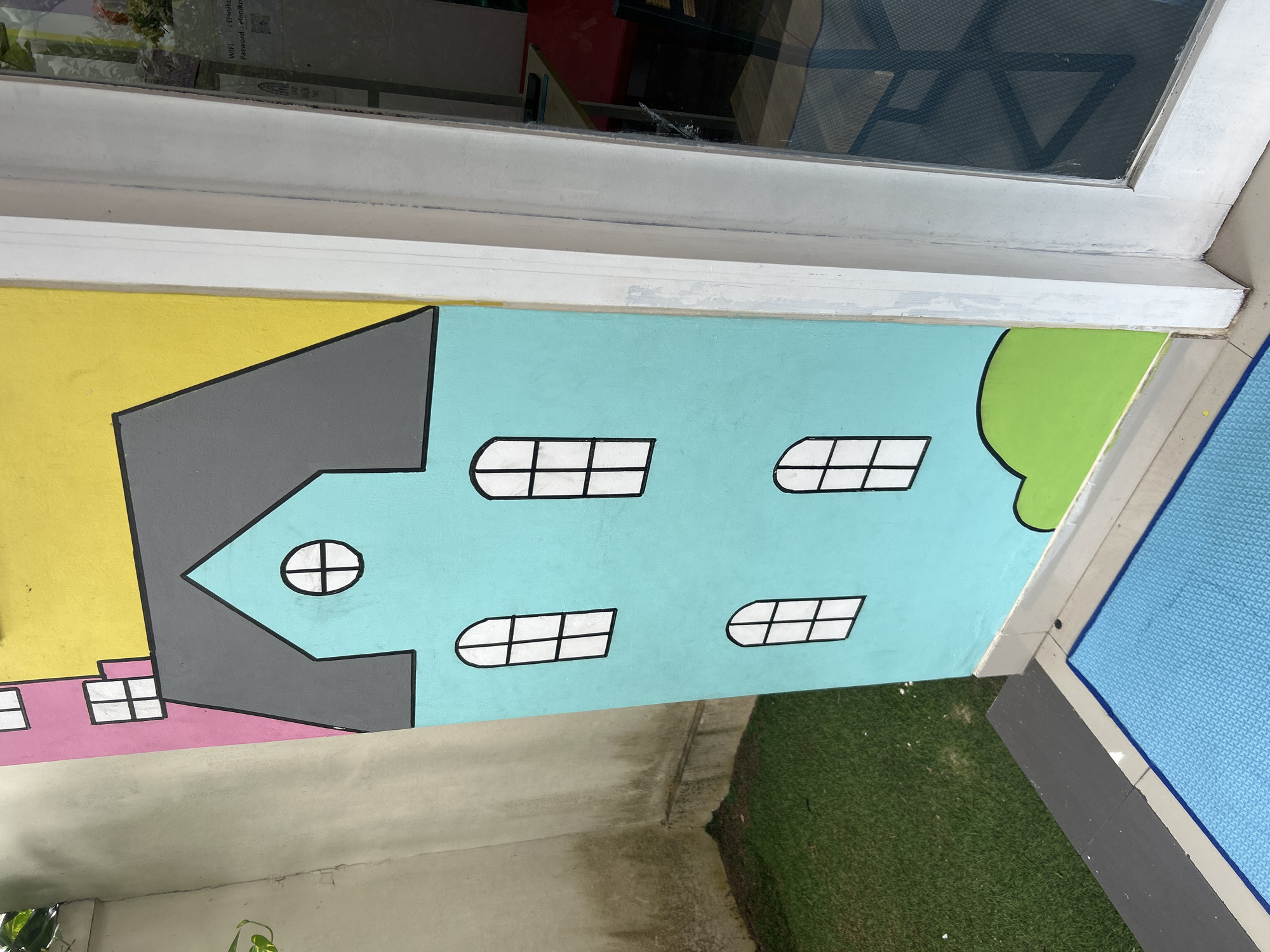An exterior view captures a vibrant mural of cartoon-like houses painted on a wall adjacent to a grassy area and a blue mat. The mural features a teal-colored house with a gray roof, adorned with black-outlined windows, two of which are circular. To the left of the teal house, a portion of a pink house with a white-outlined window is visible. The wall is further accented with a bright yellow section above the houses. To the right of the mural, a white-framed door or window with a reflective glass pane is present. The environment suggests an outdoor setting, possibly a public space or a decorative wall in Kuta, Indonesia. The focus is on the colorful artwork and its immediate surroundings.