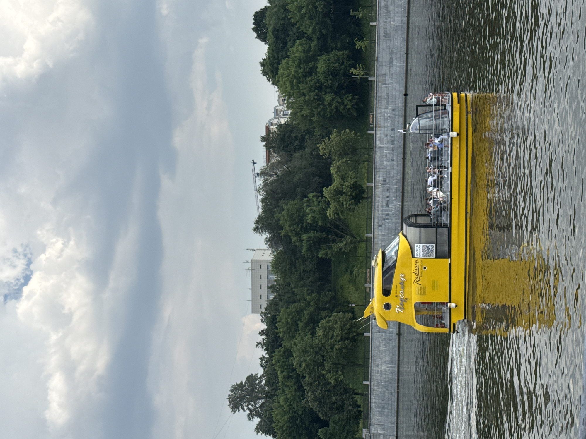 A bright yellow tourist boat with a modern, futuristic design is cruising on a calm river in Moscow, Russia. The boat has a distinctive aerodynamic shape, with a glass-enclosed cabin at the rear and an open-air seating area towards the front. Several passengers are visible inside the cabin and on the open deck, enjoying the ride. The boat is leaving a wake behind it, indicating it is in motion.
The riverbanks are lined with a stone embankment, topped by a lush green treeline. Beyond the trees, several buildings and a construction crane are visible against a cloudy sky. The overall scene suggests a pleasant day for sightseeing and enjoying the Moscow cityscape from the water. The boat is labeled with "Radisson" in prominent red lettering, indicating it is part of their river cruise services. The text below it in Cyrillic translates to "Tickets Online". The boat's name is written in Cyrillic as "Подмосковье" which translates to "Moscow Region".