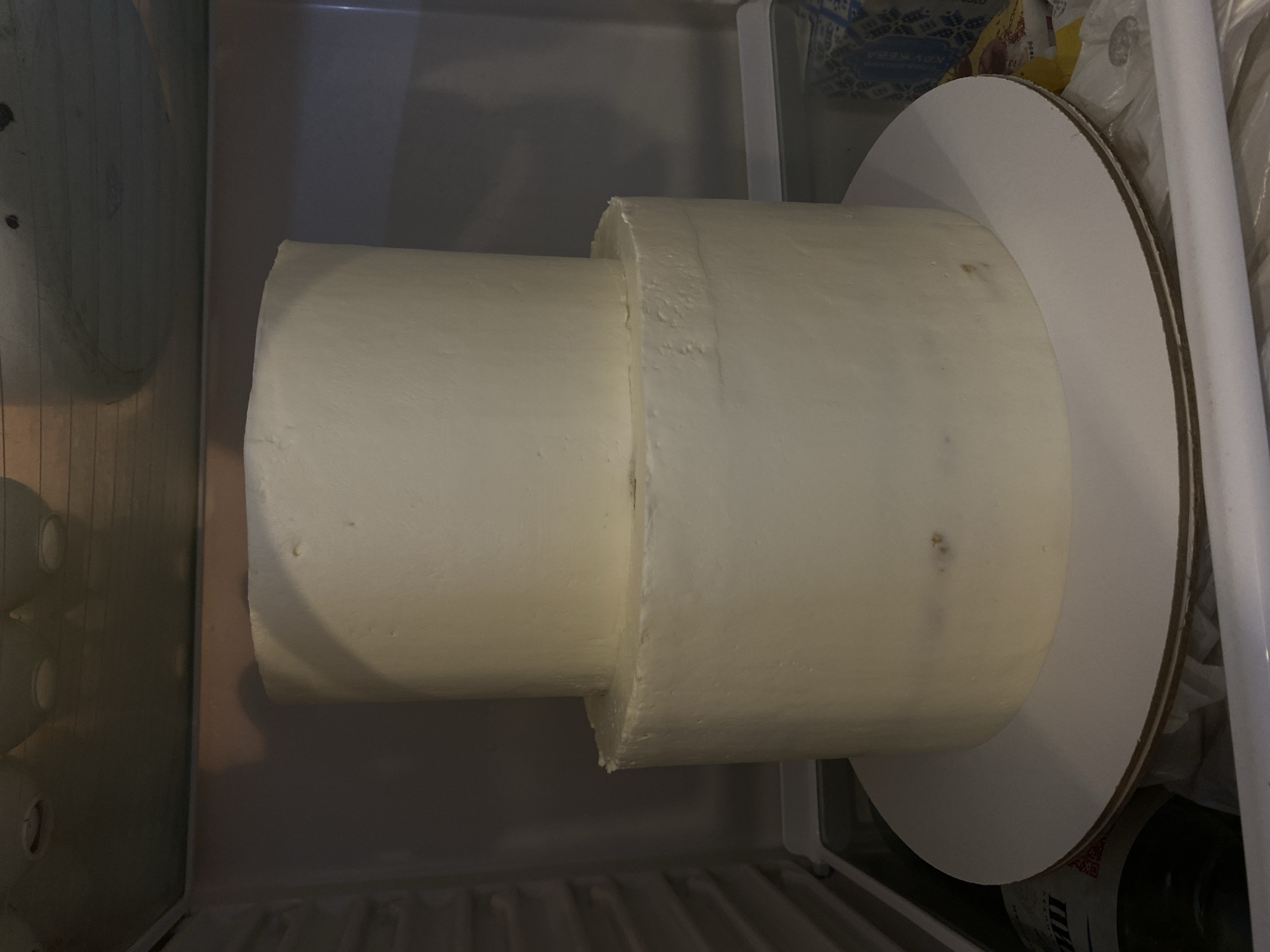 A two-tiered white frosted cake sits on a white cake board inside a refrigerator. The lower tier is larger in diameter than the upper tier. The frosting appears to be smooth with some minor imperfections. The background shows the interior of the refrigerator with shelves and possibly some food items on lower shelves. The lighting suggests the refrigerator door is open. The image does not contain any recognizable landmarks or features that would allow for identification of the city of Lomonosov, Russia.