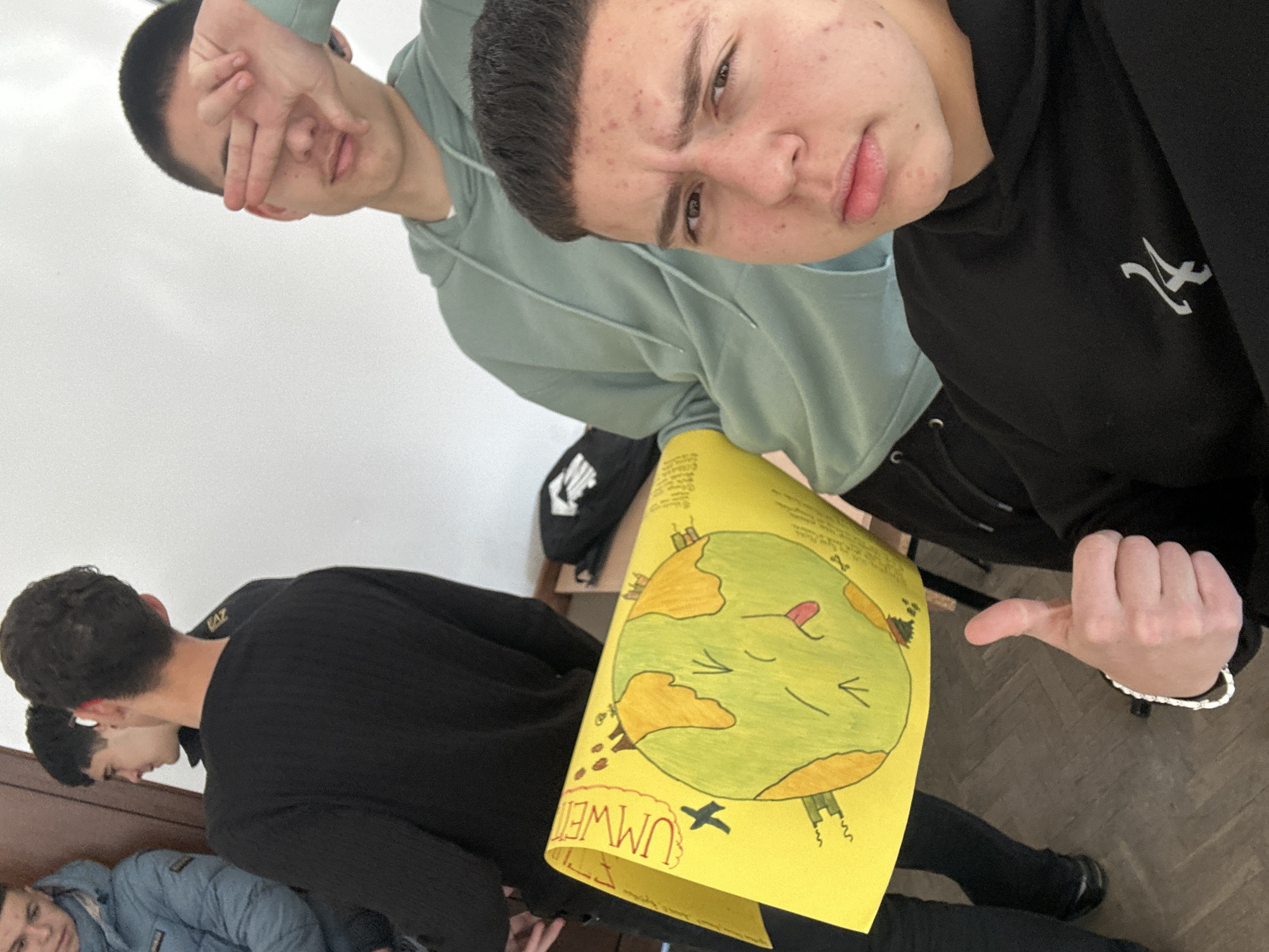 The image appears to be taken indoors, possibly in a classroom or similar setting. Three young men are visible, with two in the foreground holding a yellow poster that reads "UMWELT" and features a drawing of the Earth with cartoon-like features and imagery related to environmental concerns. One of the men is making a thumbs-down gesture towards the poster. The third man is partially visible in the background and appears to be looking in a different direction. Given the caption provided, it can be inferred that this scene is taking place in Zlatograd, Bulgaria.