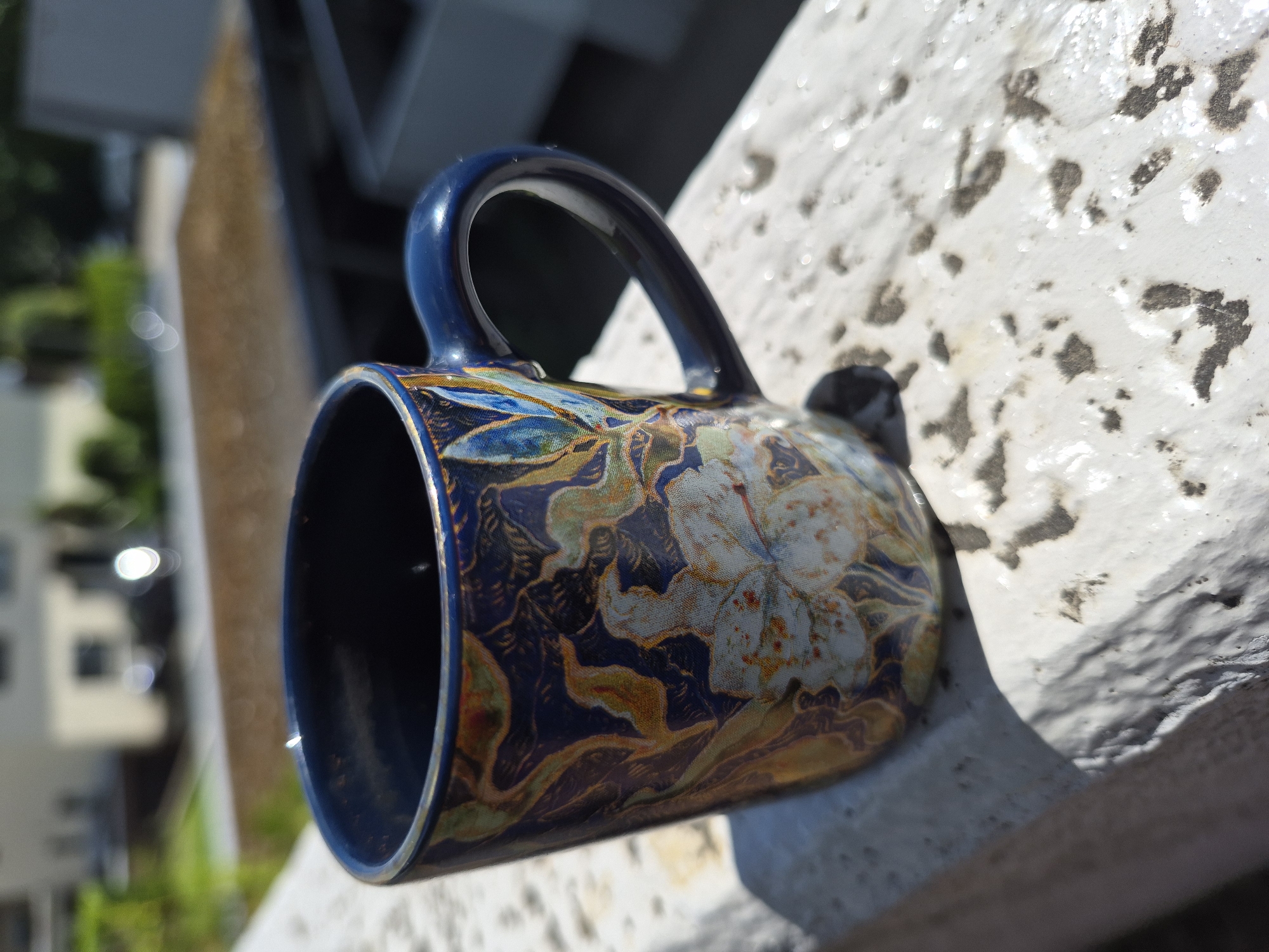 A blue mug with a floral design sits on a white ledge. The mug is dark blue with intricate patterns of light blue, gold, and white flowers and leaves. The handle is dark blue and curves outwards. The white ledge appears to be weathered, with some dark spots and texture. In the background, out of focus, are buildings and greenery, suggesting an outdoor urban or suburban setting. The lighting indicates it is daytime, with sunlight casting shadows. There are no people or animals visible.