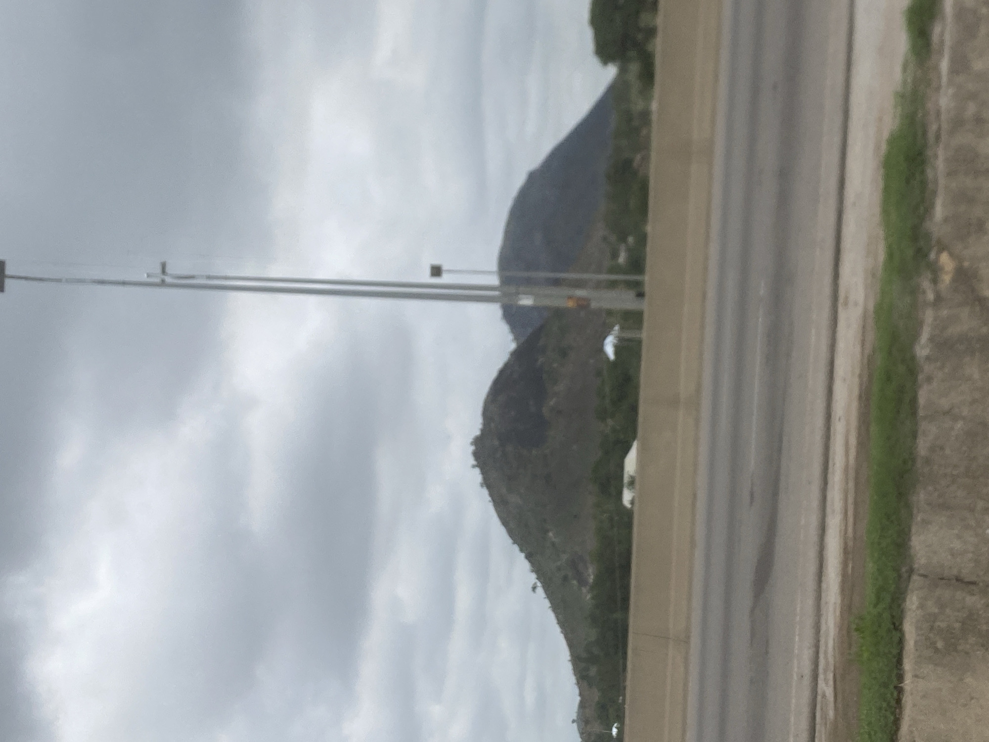 The image presents a landscape featuring mountains, an overcast sky, and a section of a road with utility poles. The scene is dominated by a light grey, cloudy sky that fills the upper portion of the frame, suggesting an overcast day.
In the middle ground, two prominent, dark-colored mountains or large hills rise, their slopes appearing rugged and covered with sparse, dark green vegetation. Their irregular peaks are silhouetted against the cloudy sky.
On the right side of the image, a multi-lane paved road or highway is visible, bordered by a concrete barrier. A thin strip of green grass grows alongside this barrier, adjacent to another concrete structure or embankment. Several tall, slender, metallic utility poles or street light poles stand in the foreground, extending upwards from near the road level. Wires or cables are strung horizontally between these poles.
A small, light-colored vehicle, possibly a white van or truck, is partially visible on the road behind the concrete barrier, positioned towards the base of one of the mountains. There is no discernible text in the image. The setting appears to be an outdoor, daytime scene, likely in an area like Gosa, Nigeria, characterized by both natural topography and human infrastructure.