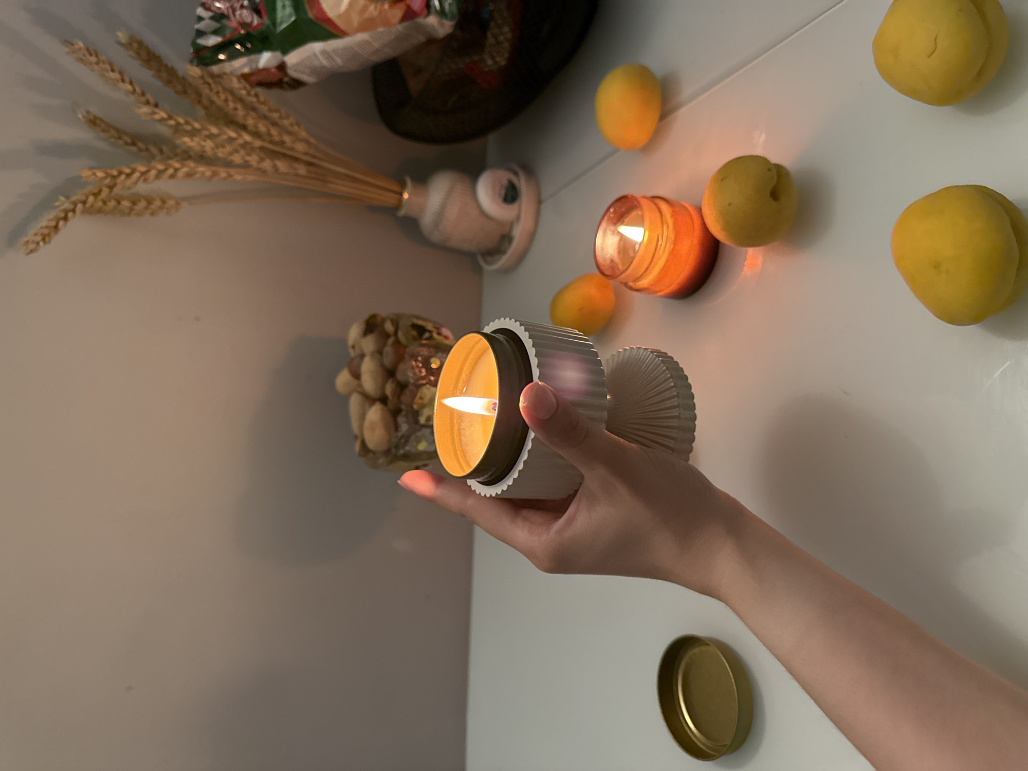 A person's hand is holding a lit candle in a decorative white holder. The candle has a bright orange flame. Nearby on a white surface are several yellow fruits, possibly apricots or small lemons, and another smaller lit candle in an amber glass jar. In the background, a vase with wheat stalks stands next to a bowl of nuts and a portion of a green and white bag. The lighting is dim, with the candles providing the main illumination. The scene appears to be indoors on a tabletop.