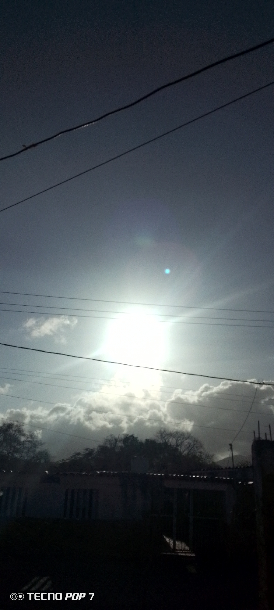 A low-angle shot shows a bright sun obscured by clouds, with power lines crossing the sky. Below, trees and a building appear silhouetted, likely in Altagracia, Venezuela. A "Tecno Pop 7" watermark is at the bottom.