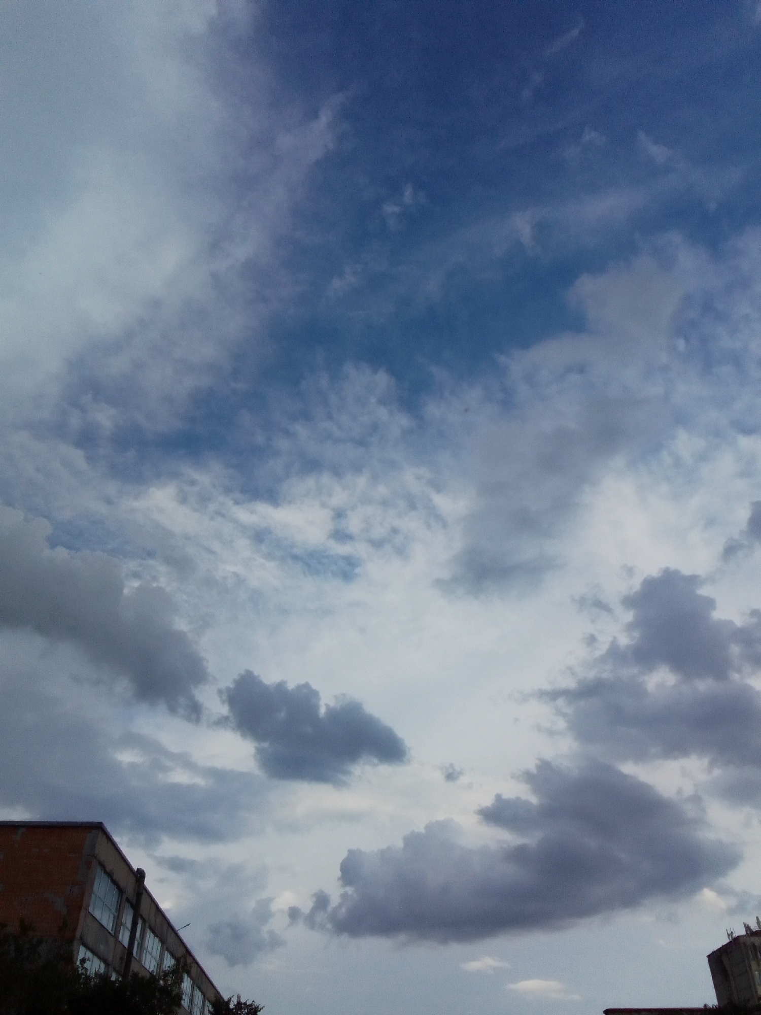 The image shows a cloudy sky above some buildings. On the left side, a red brick building with many windows is visible, partially covered by trees. The sky is a mix of blue and grey clouds, with some darker, thicker clouds towards the bottom and lighter, wispy clouds higher up. The overall lighting suggests it might be daytime, possibly overcast or late afternoon. No people are visible in the scene. The image is a photograph taken from a low angle, looking upwards towards the sky.