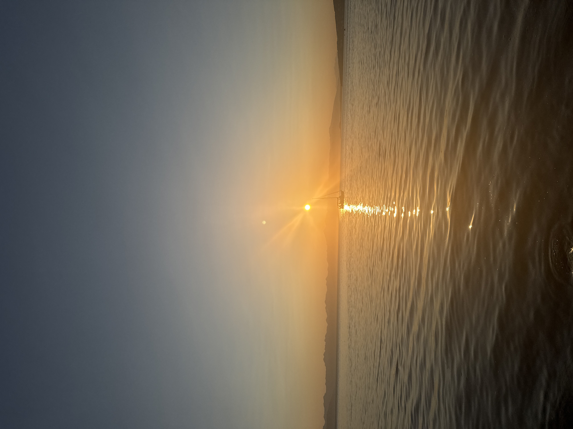 A vertical, eye-level shot captures a serene seascape at sunset. The sun, a brilliant orb with visible rays, sits low on the horizon, casting a golden glow that reflects vibrantly on the water's surface. The sky transitions from a pale blue at the top to a warm orange near the horizon, a gradient intensified by the sunset. A sailboat with its mast visible is silhouetted against the bright sun. In the distance, a faint line of mountains borders the water. The water itself is calm, with subtle ripples creating a textured surface. The scene is devoid of any people or specific landmarks that would pinpoint it to Petropavlovsk-Kamchatsky, Russia.