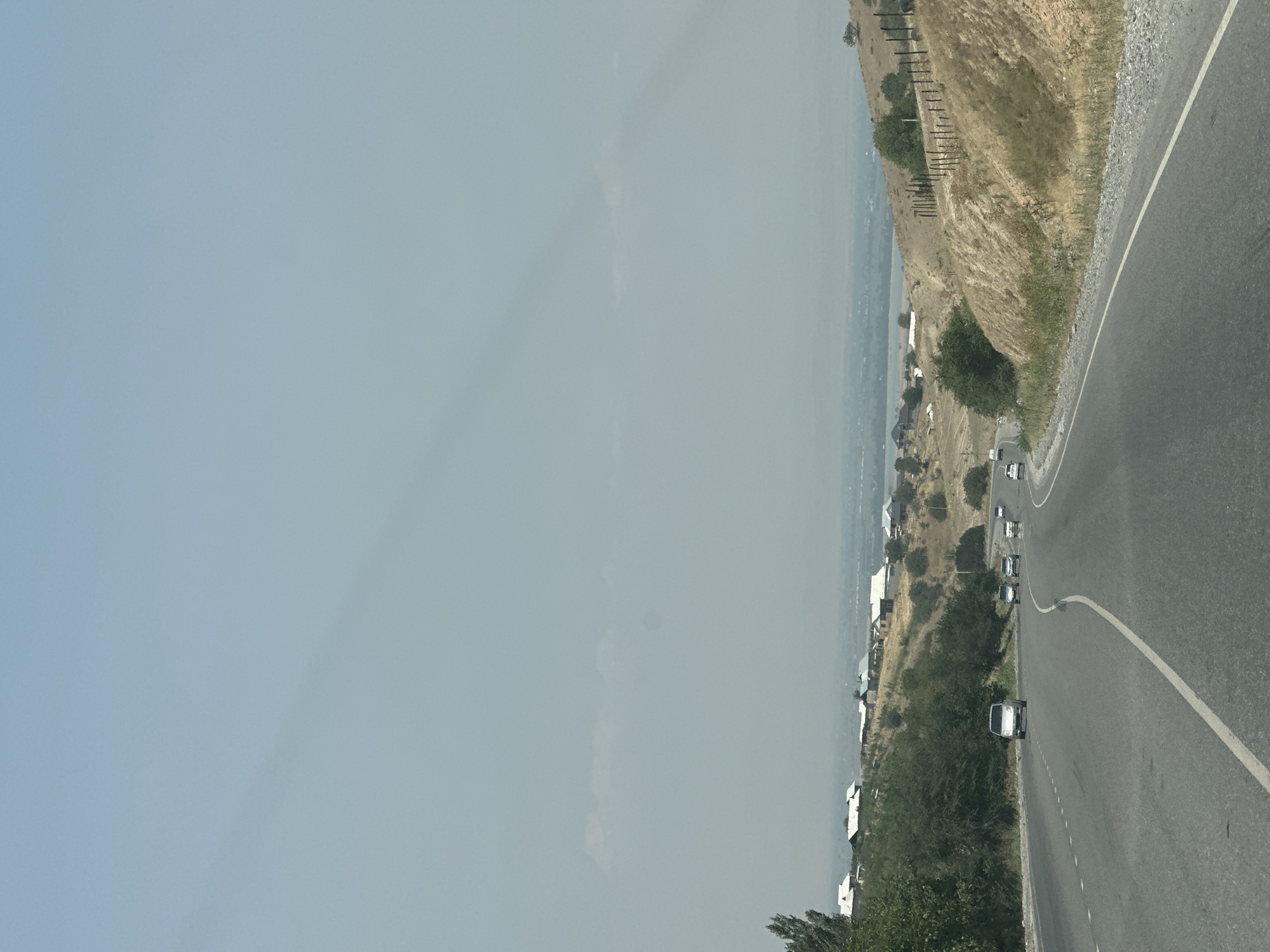 A curved asphalt road with a white dashed line runs through a rural landscape under a hazy sky. The road is bordered by dry, grassy hills on the right and a green, tree-covered slope on the left. Several cars are visible on the road, moving in the same direction as the viewer.
In the background, a collection of houses with white roofs are clustered on the hillside. Beyond the houses, a body of water, possibly a lake or reservoir, is visible in the distance, surrounded by a hazy, flat expanse that suggests a valley or plain. A fence made of wooden posts runs along the top of the hill on the right.
The scene appears to be a roadside view on a somewhat overcast or dusty day, capturing a glimpse of the outskirts of Suzak, Kyrgyzstan, where a semi-arid environment meets human settlement and infrastructure.