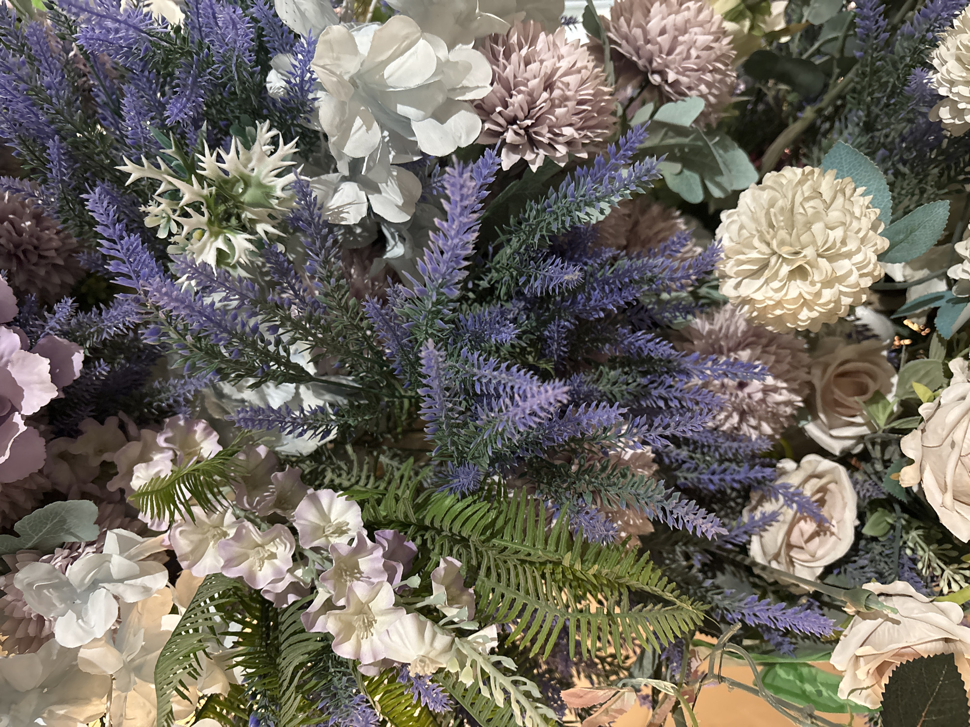 This is a close-up, eye-level photograph of a dense floral arrangement. The arrangement is composed of a variety of artificial flowers and foliage in shades of purple, white, cream, and green. Prominently featured are clusters of lavender-like purple flowers, large white and light pink blossoms, and delicate ferns. The composition is rich and textural, with many layers and overlapping elements. The lighting appears to be artificial, casting soft shadows and highlighting the different textures of the flowers. There are no people or animals visible in the image. The background is out of focus, suggesting the arrangement is the sole subject of the photograph. There is no information in the image to suggest a location in Shymkent, Kazakhstan.